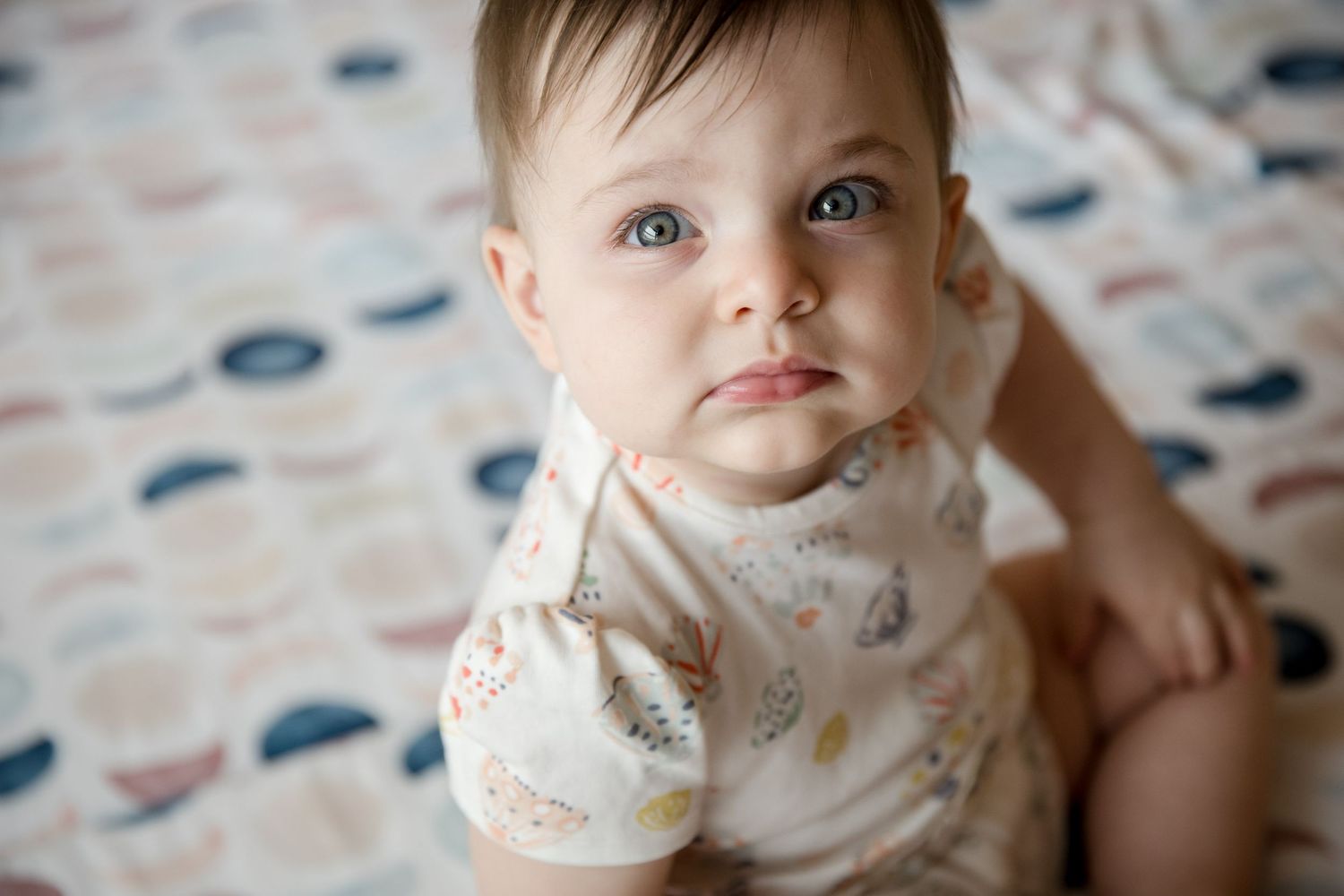 Adorable baby with blue eyes sitting on a patterned blanket wearing a white floral onesie, looking up curiously.