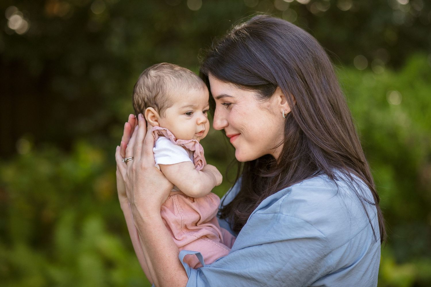 Loving mother with long dark hair tenderly holds her baby girl outdoors with green foliage background.