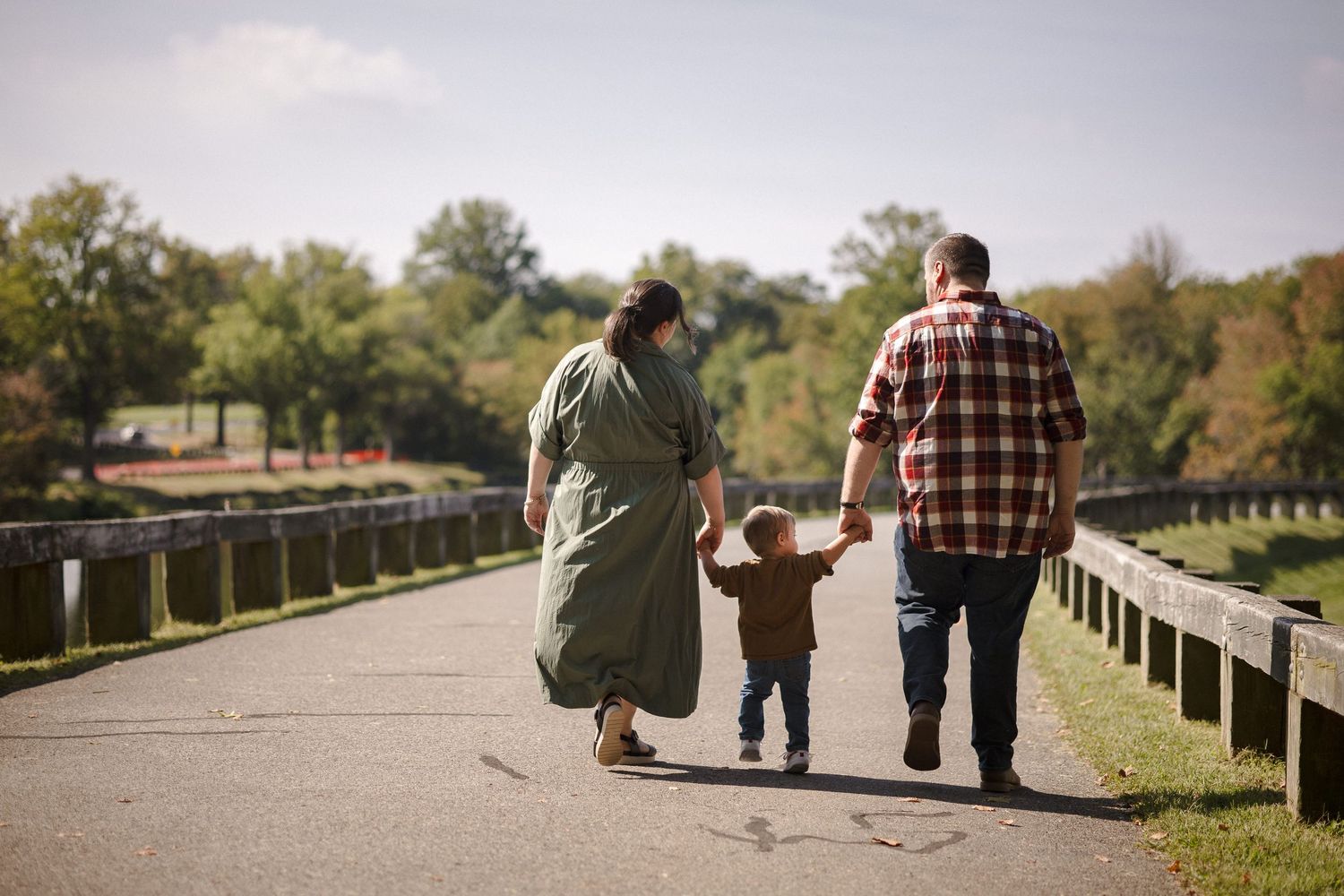 Family of three walking hand-in-hand down a scenic park path surrounded by lush green trees and a wooden fence.