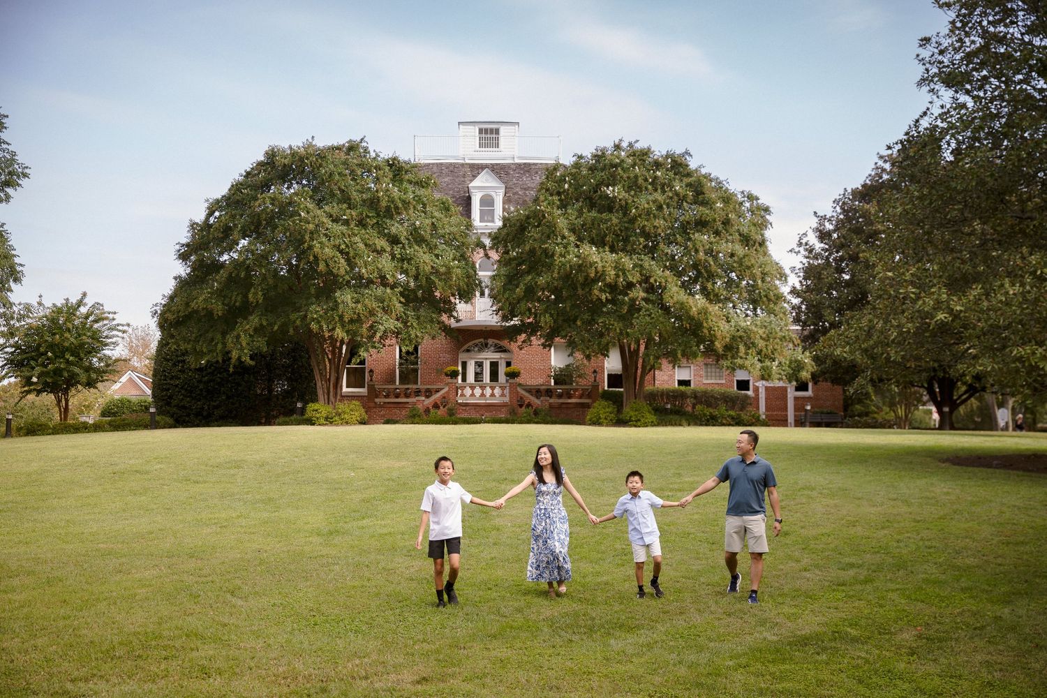 Family of four holding hands walking on green lawn in front of large brick estate home surrounded by trees.