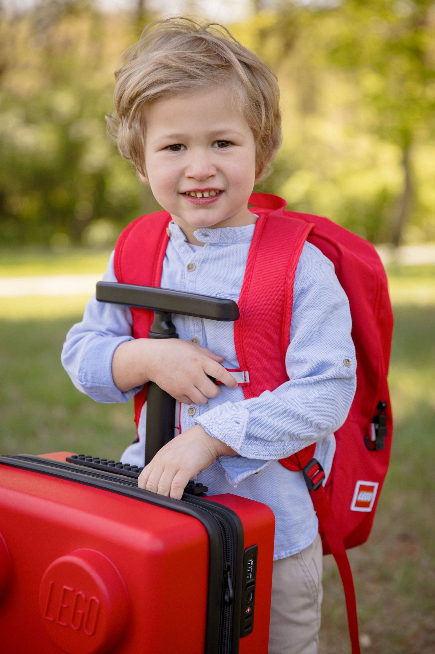 Smiling toddler boy with red LEGO backpack and suitcase outdoors in a park setting.