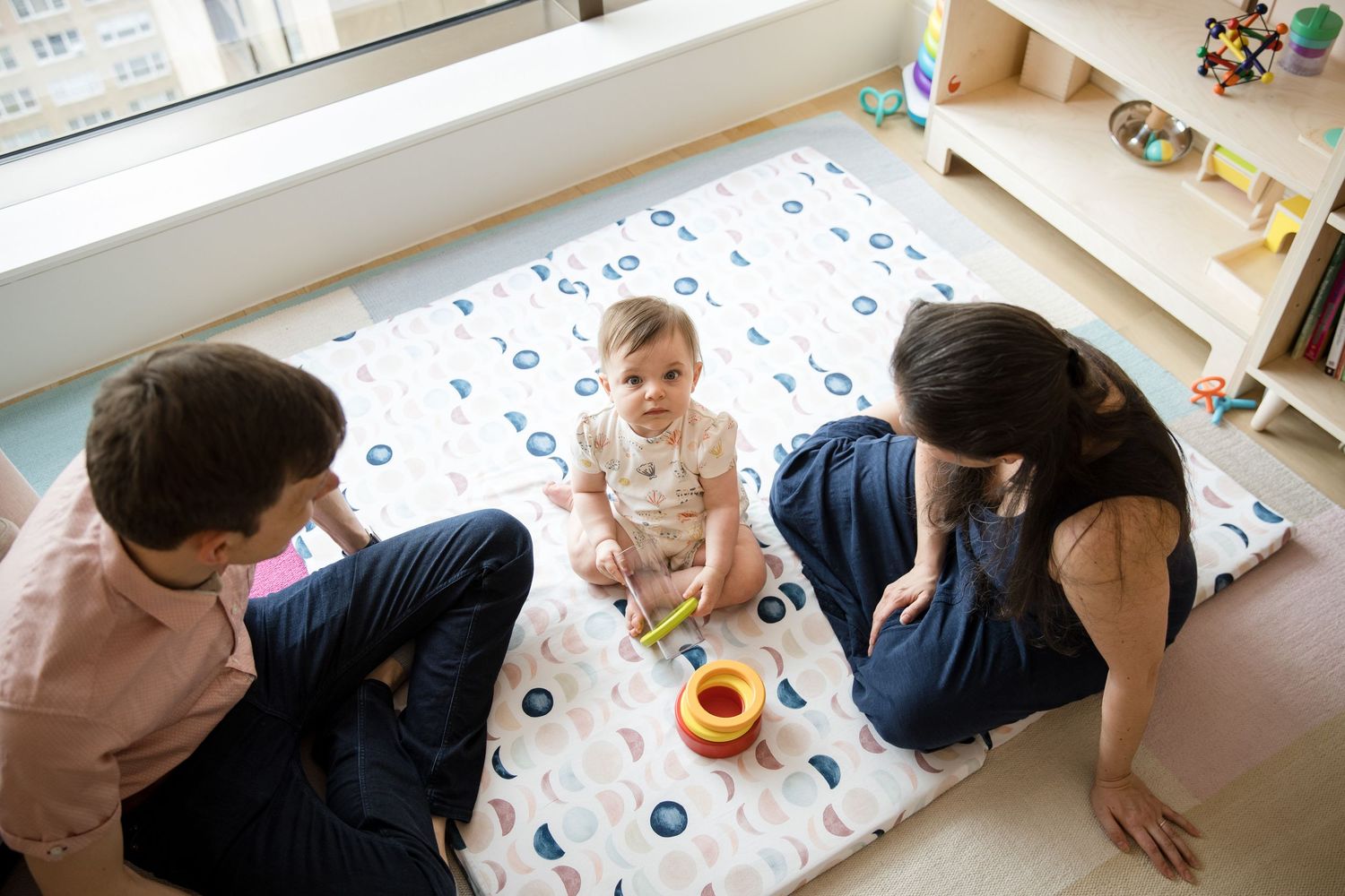 Two adults sit on a patterned play mat with a baby playing with colorful stacking toys in a bright nursery room.