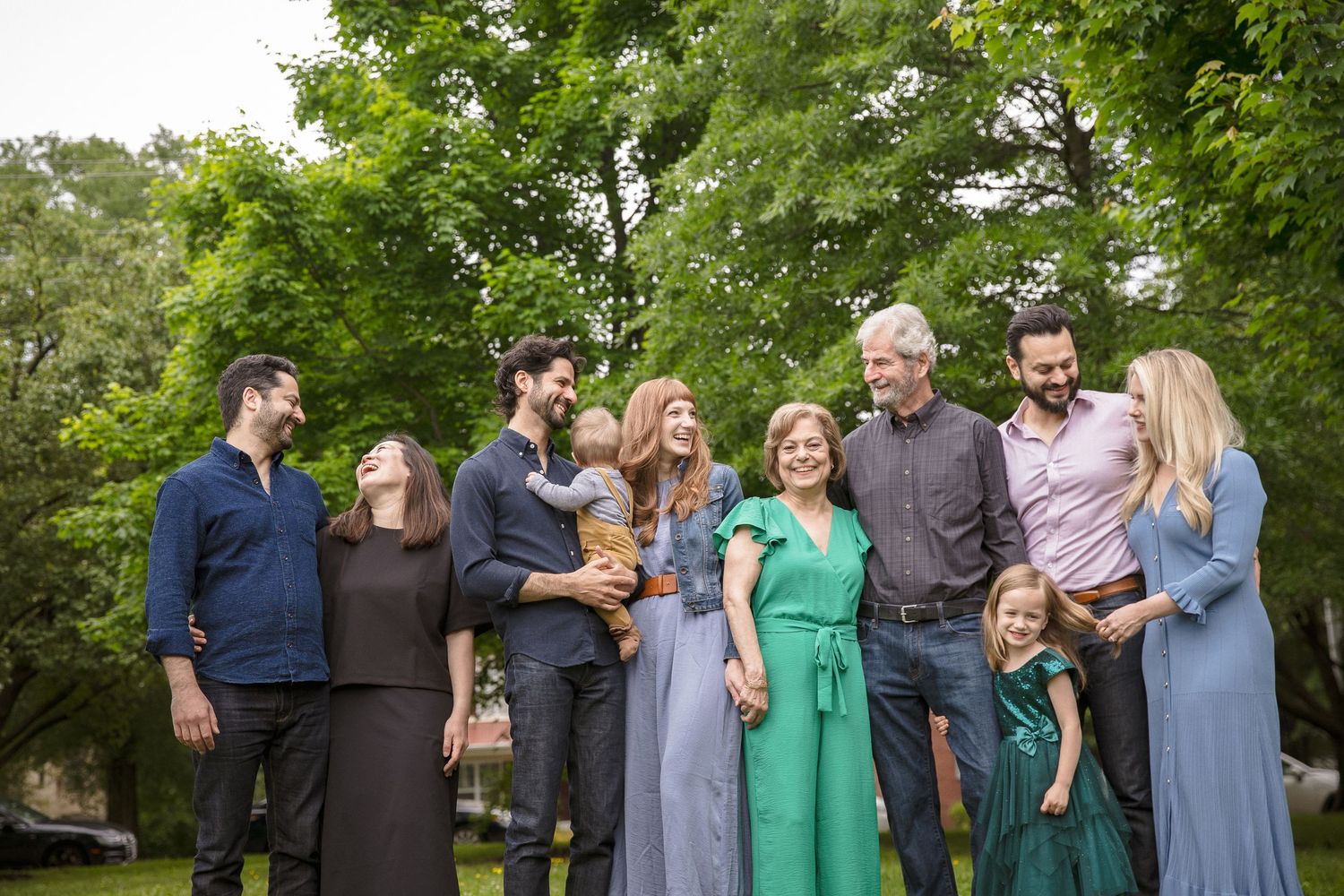 Large multigenerational family group laughing and smiling together outdoors under lush green trees.