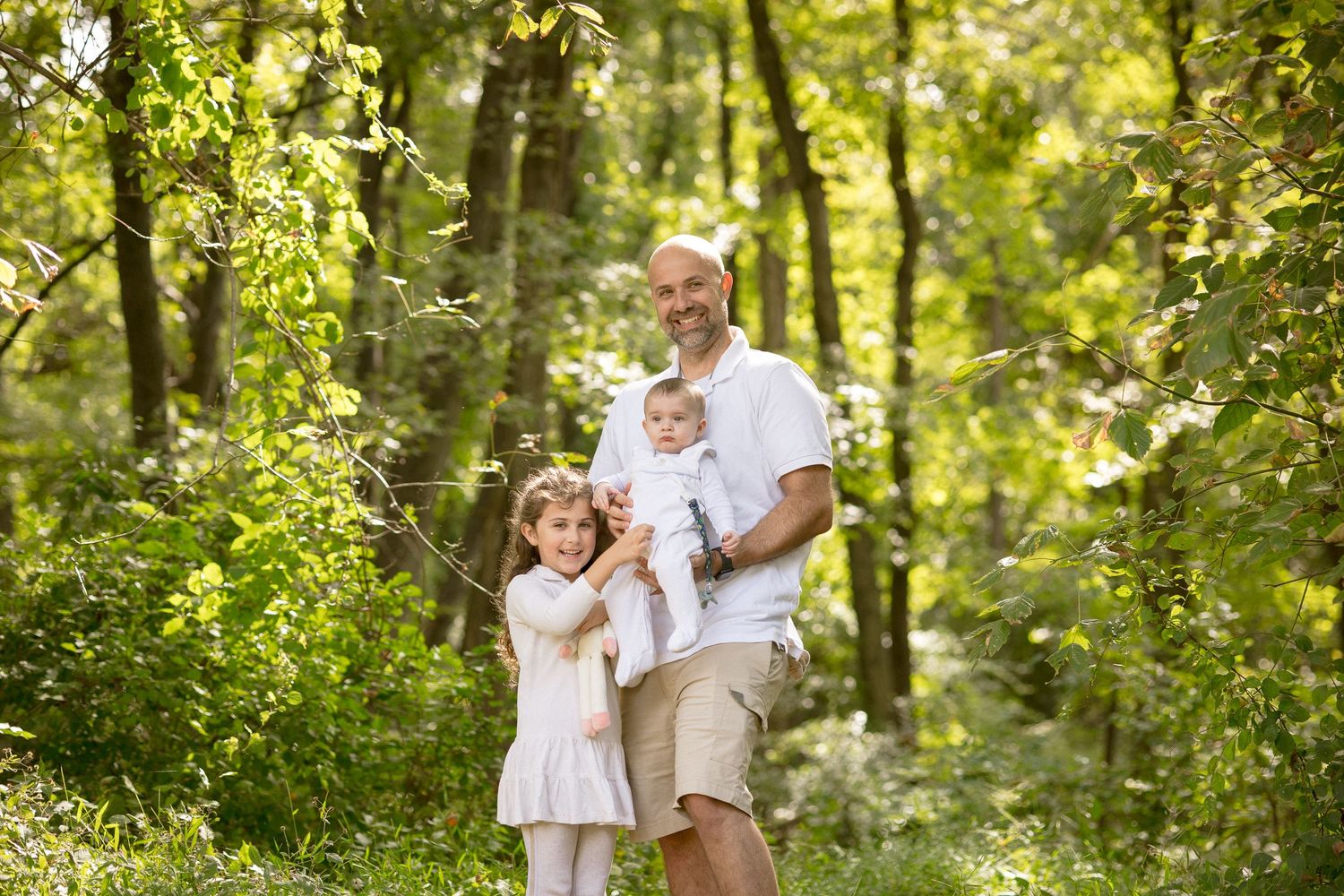 A father smiles holding a baby and standing with his young daughter in a lush green forest.