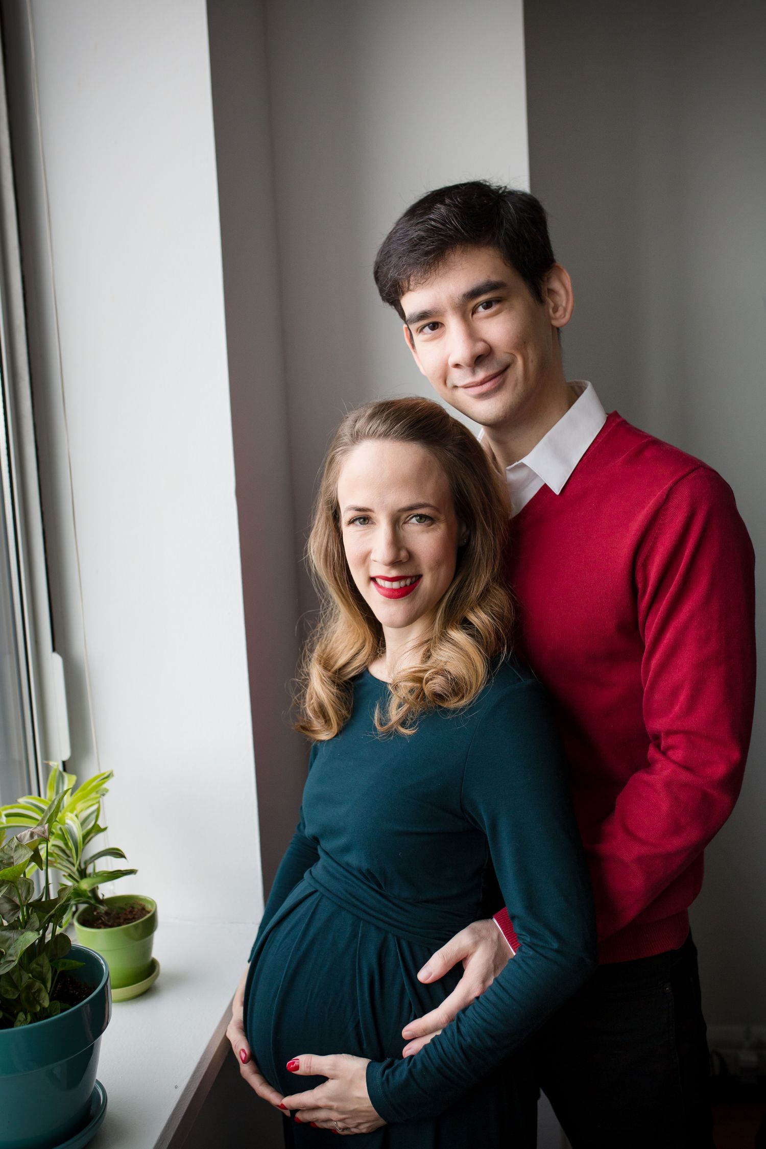Pregnant couple posing by window, man in red sweater embracing woman in teal dress while holding her baby bump.