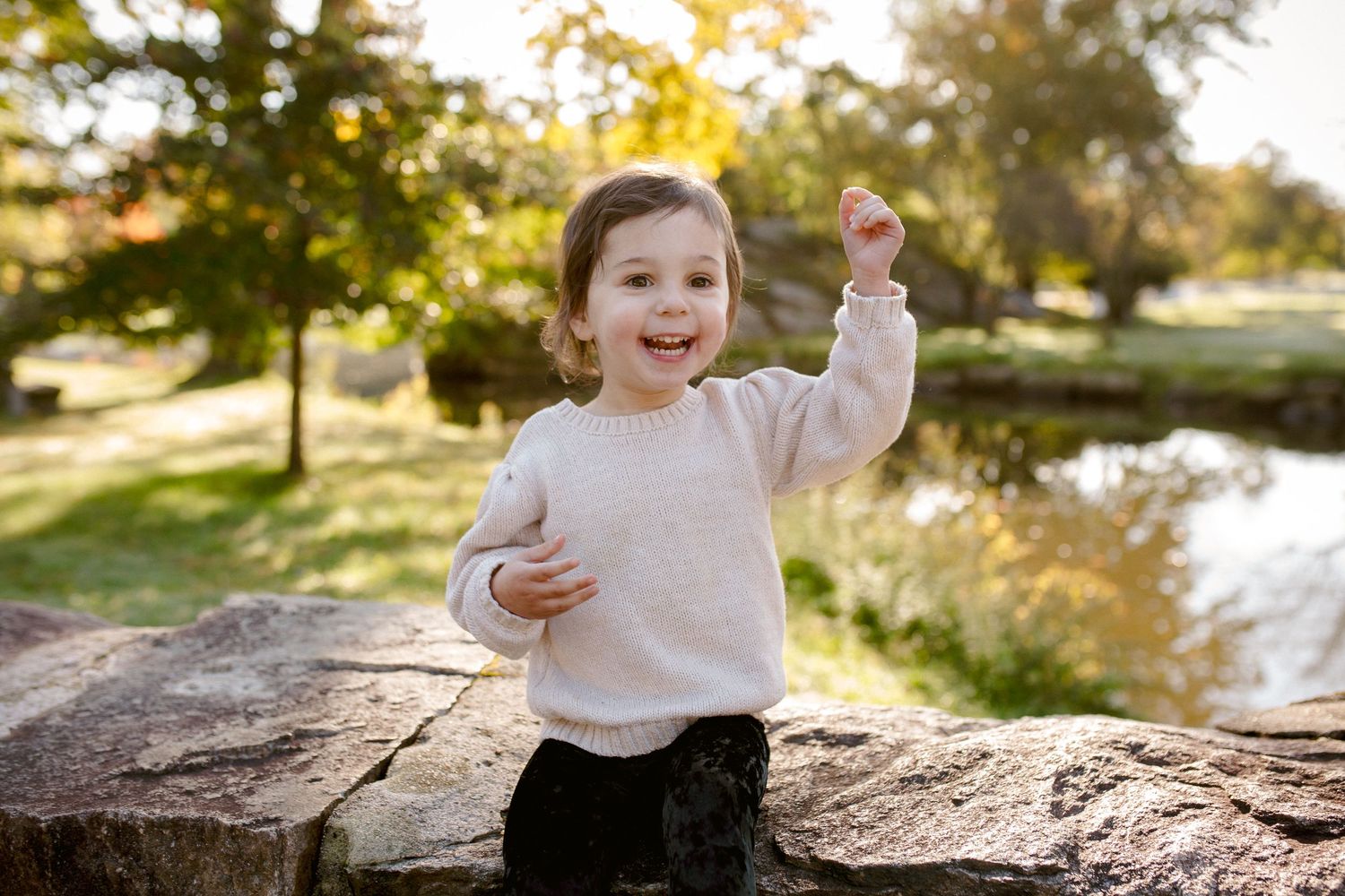 Happy toddler in white sweater raising arm joyfully while standing on rocks in a sunny park.