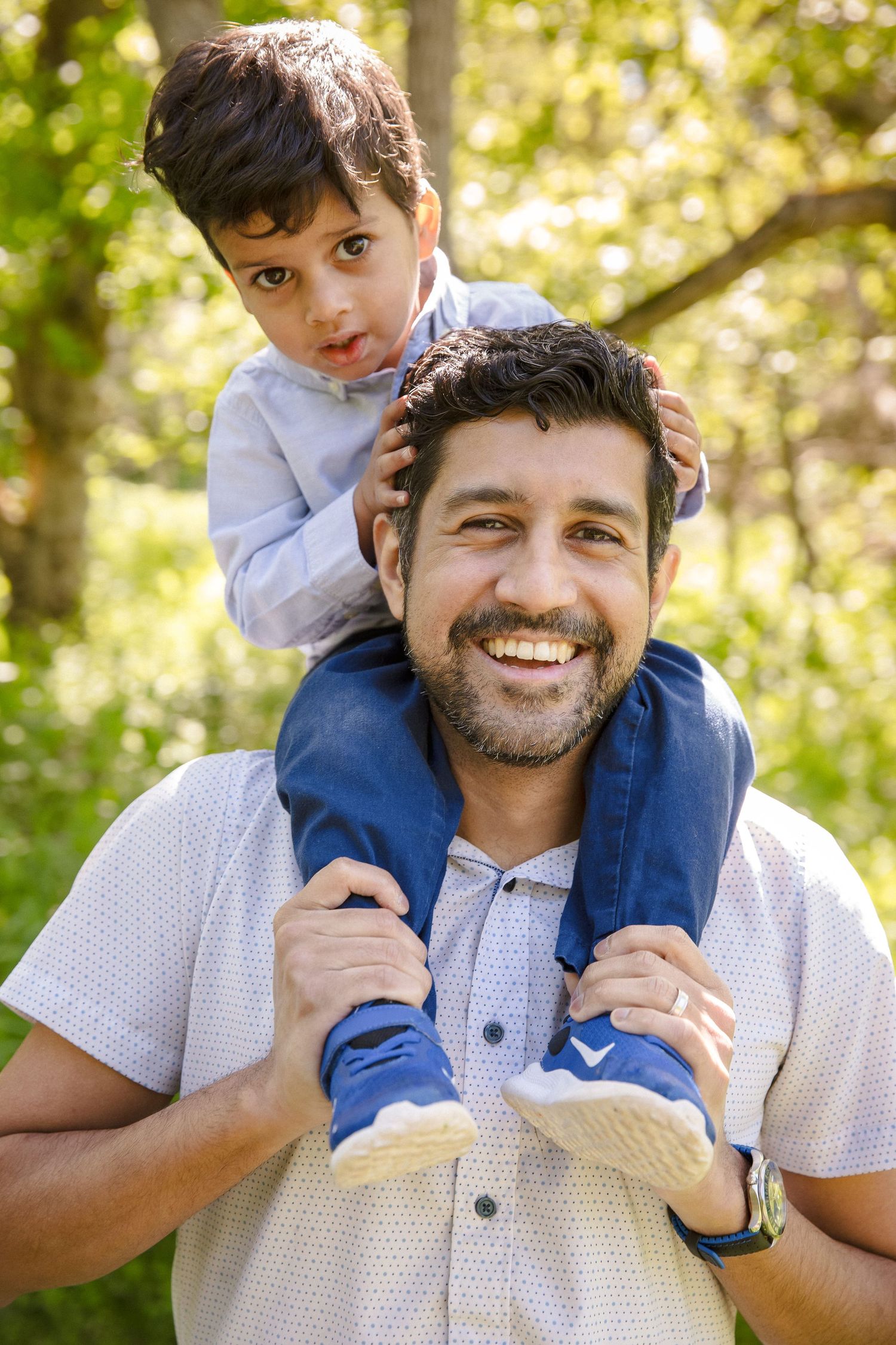 Smiling father carries young son on his shoulders in a sunny green outdoor setting.