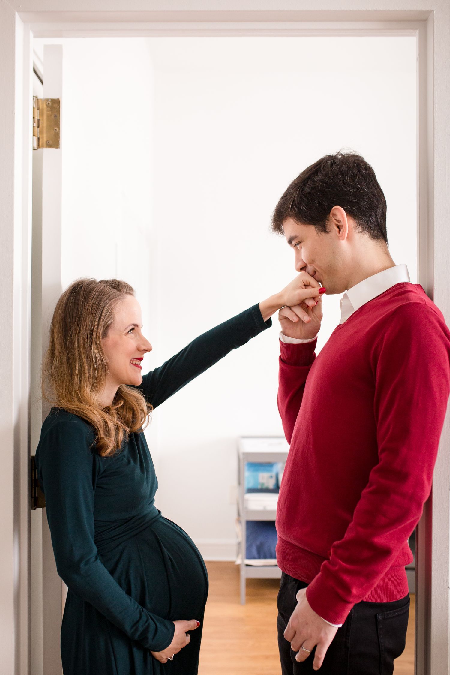 A man in a red sweater kisses the hand of a smiling pregnant woman in a green dress in a doorway.