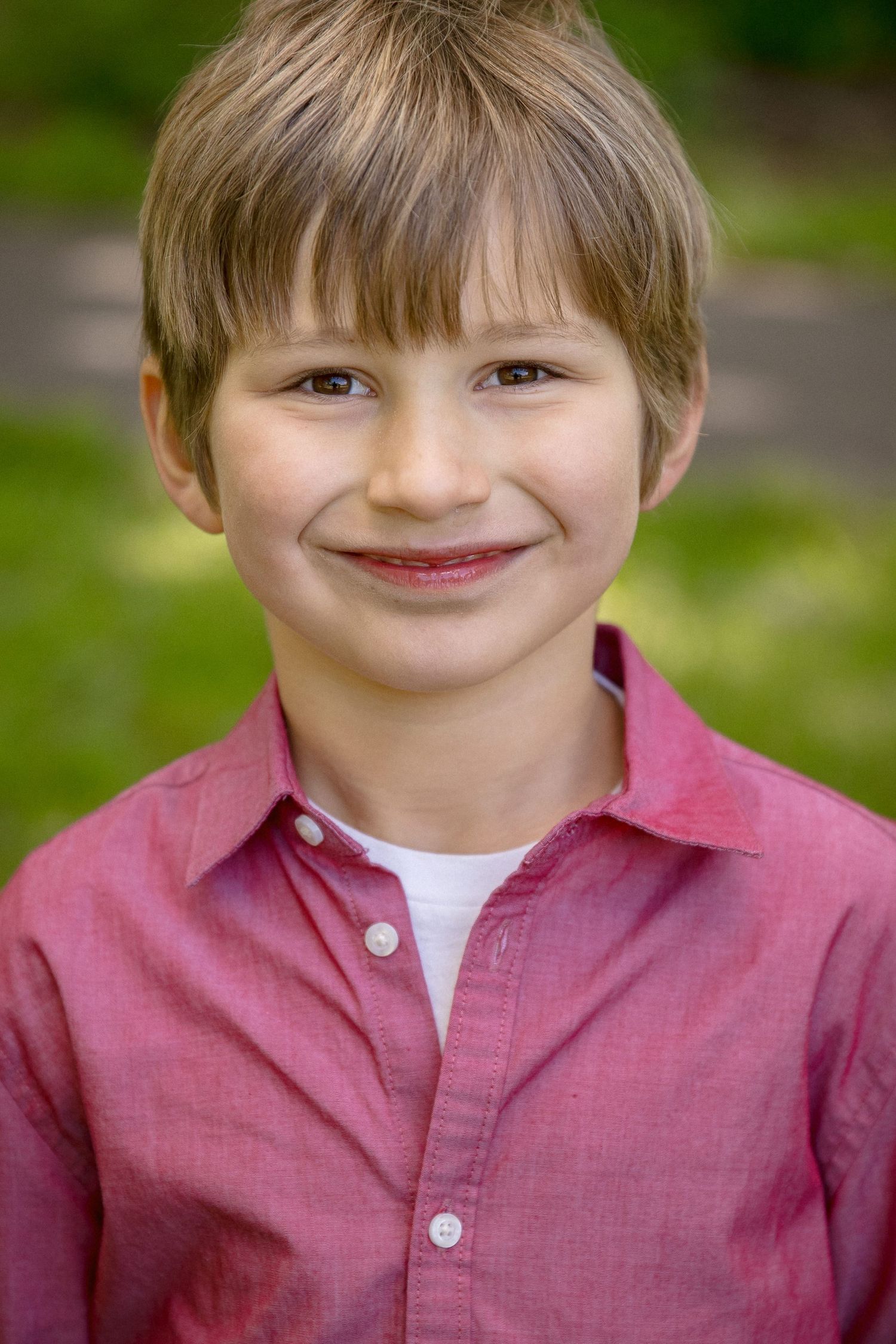 Smiling young boy with blonde hair wearing a red button-up shirt outdoors with green foliage background.