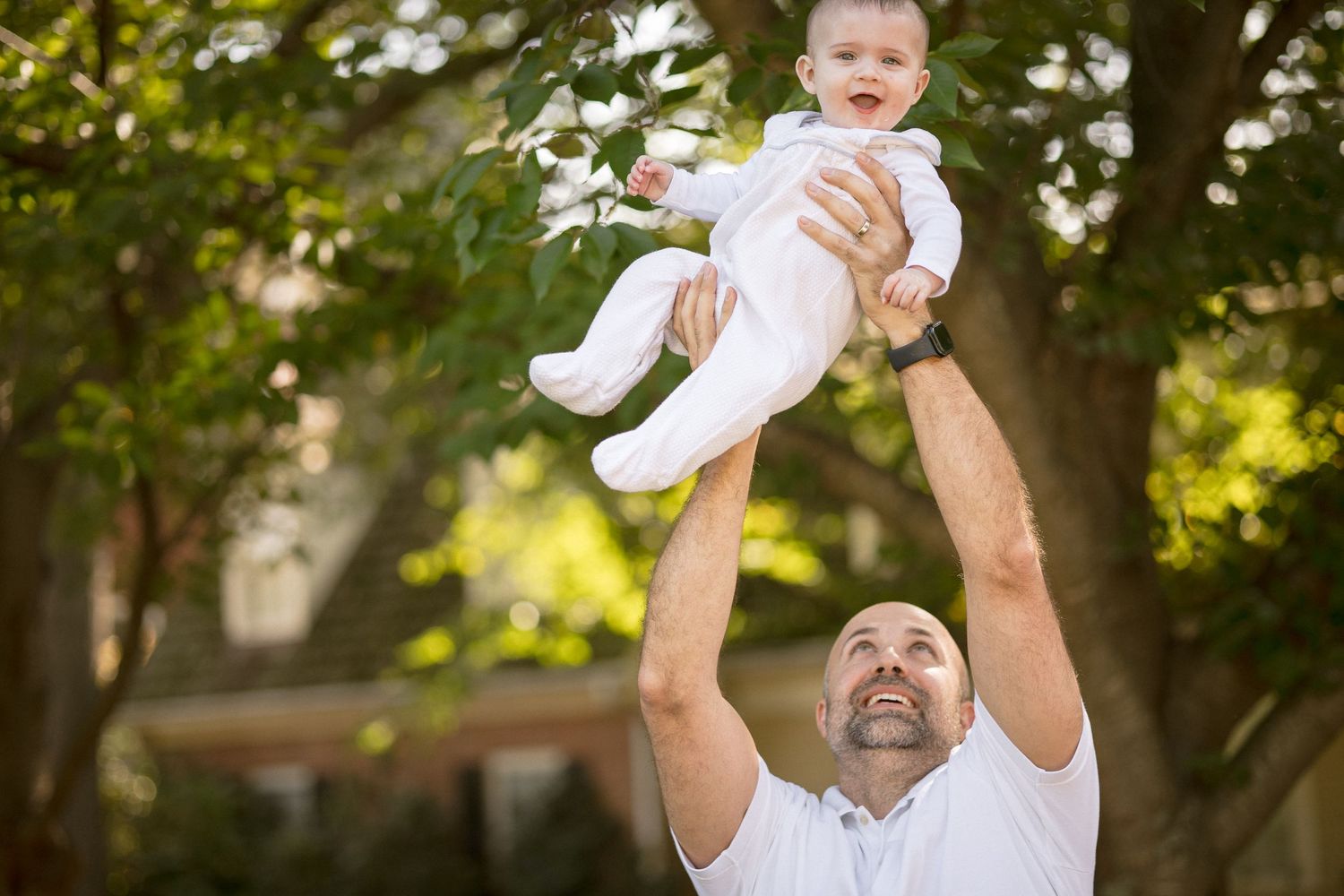 Father joyfully lifting laughing baby high in the air outdoors under green trees.