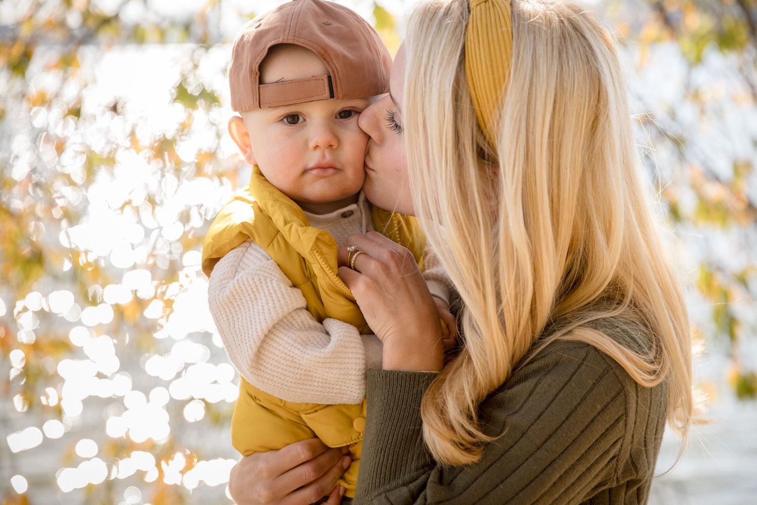 Blonde mother tenderly kisses her baby dressed in yellow outdoors in fall.