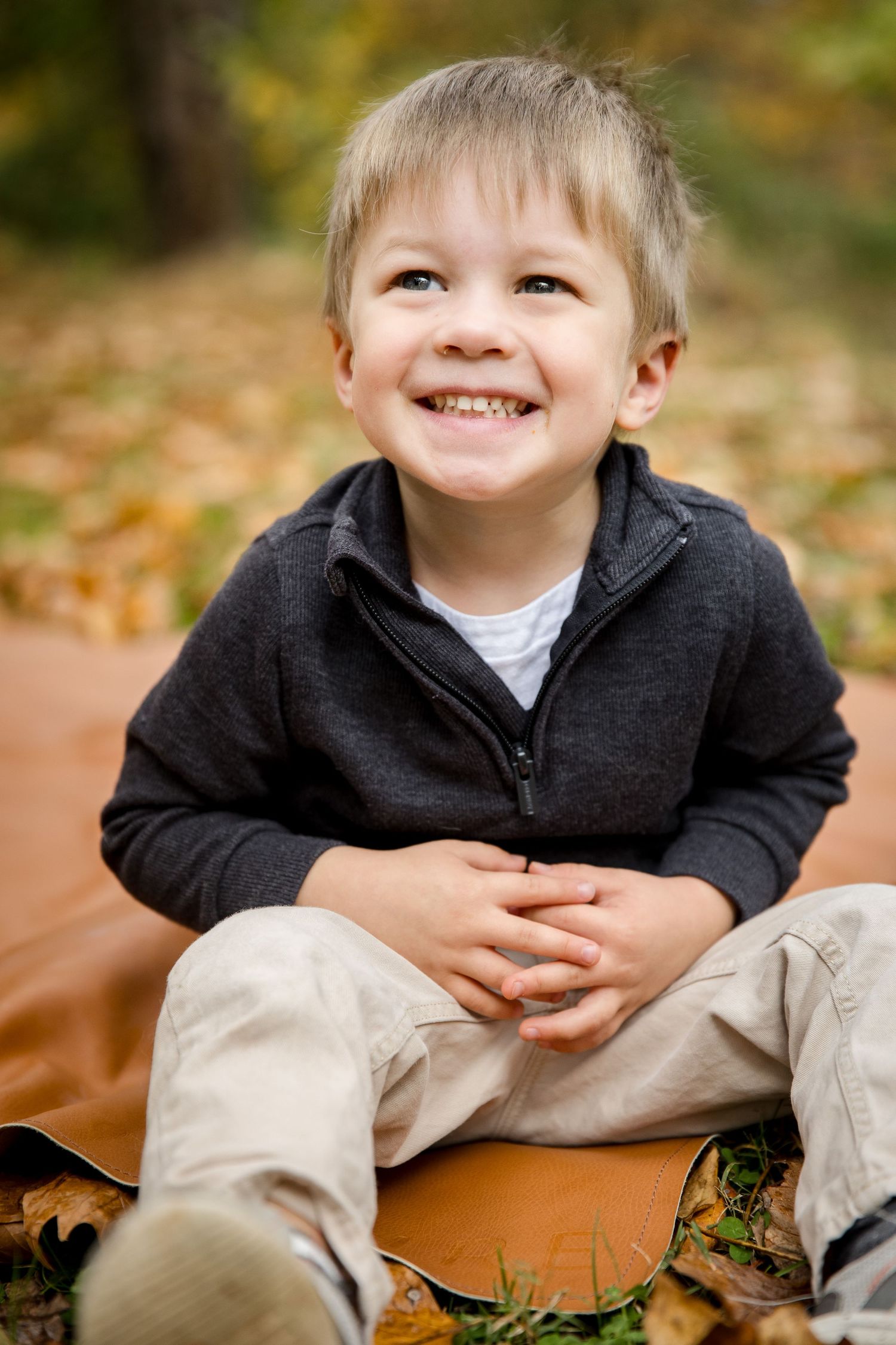 Happy young boy smiling while sitting on a blanket surrounded by autumn leaves in a park.