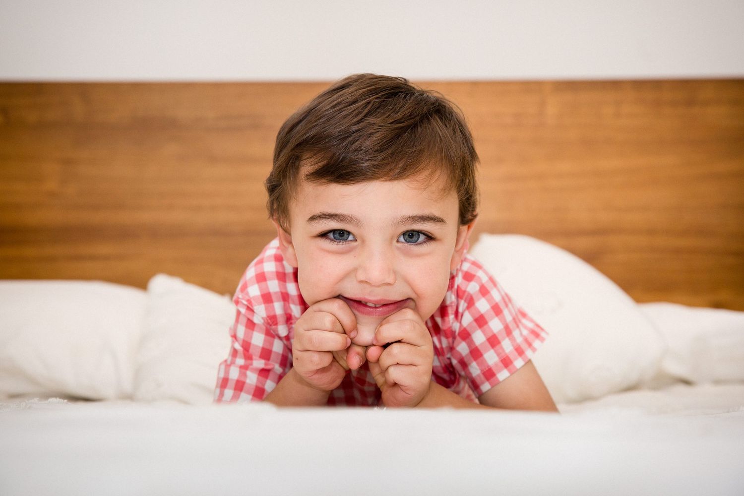 Cute toddler boy lying on white bed smiling with chin on hands.