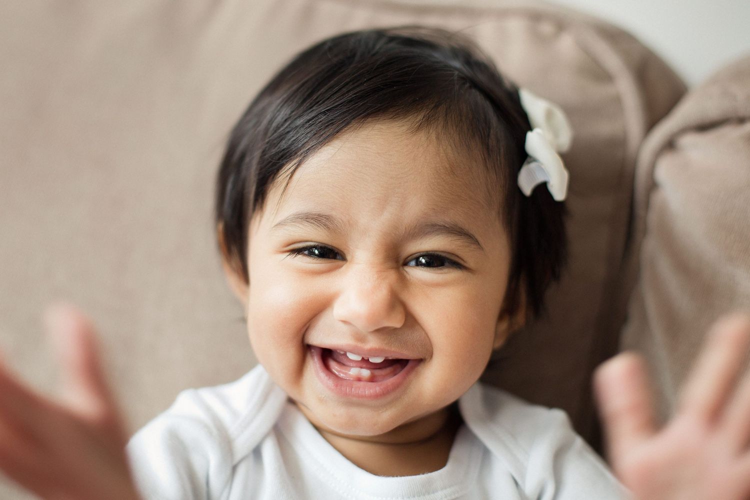 Adorable baby girl with hair bow laughing and showing tiny teeth in a joyful close-up portrait.