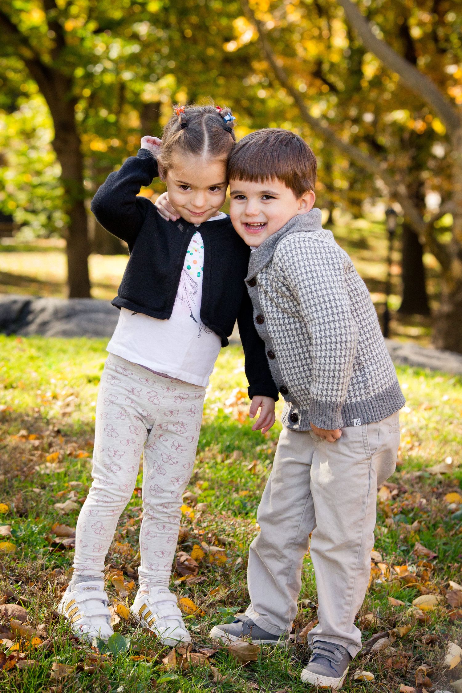 Two young children hugging and smiling in autumn park surrounded by golden fallen leaves.