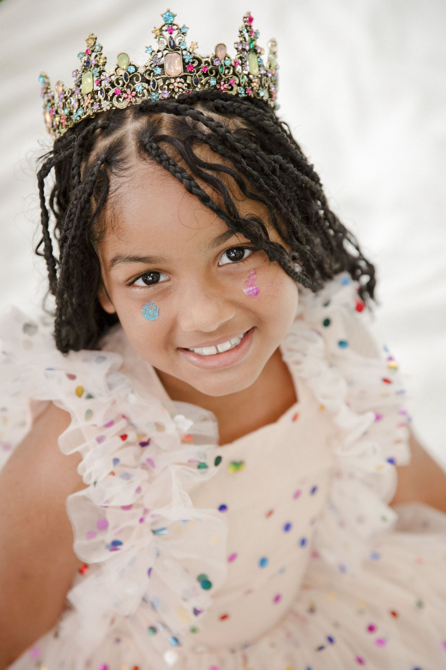 Young girl wearing a jeweled crown and sparkly white dress with glitter on her cheeks.
