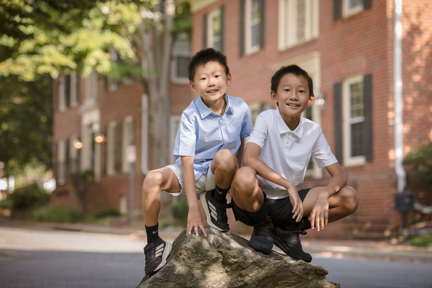 Two young boys in school uniforms squat on a rock outdoors near a brick building, smiling at the camera.