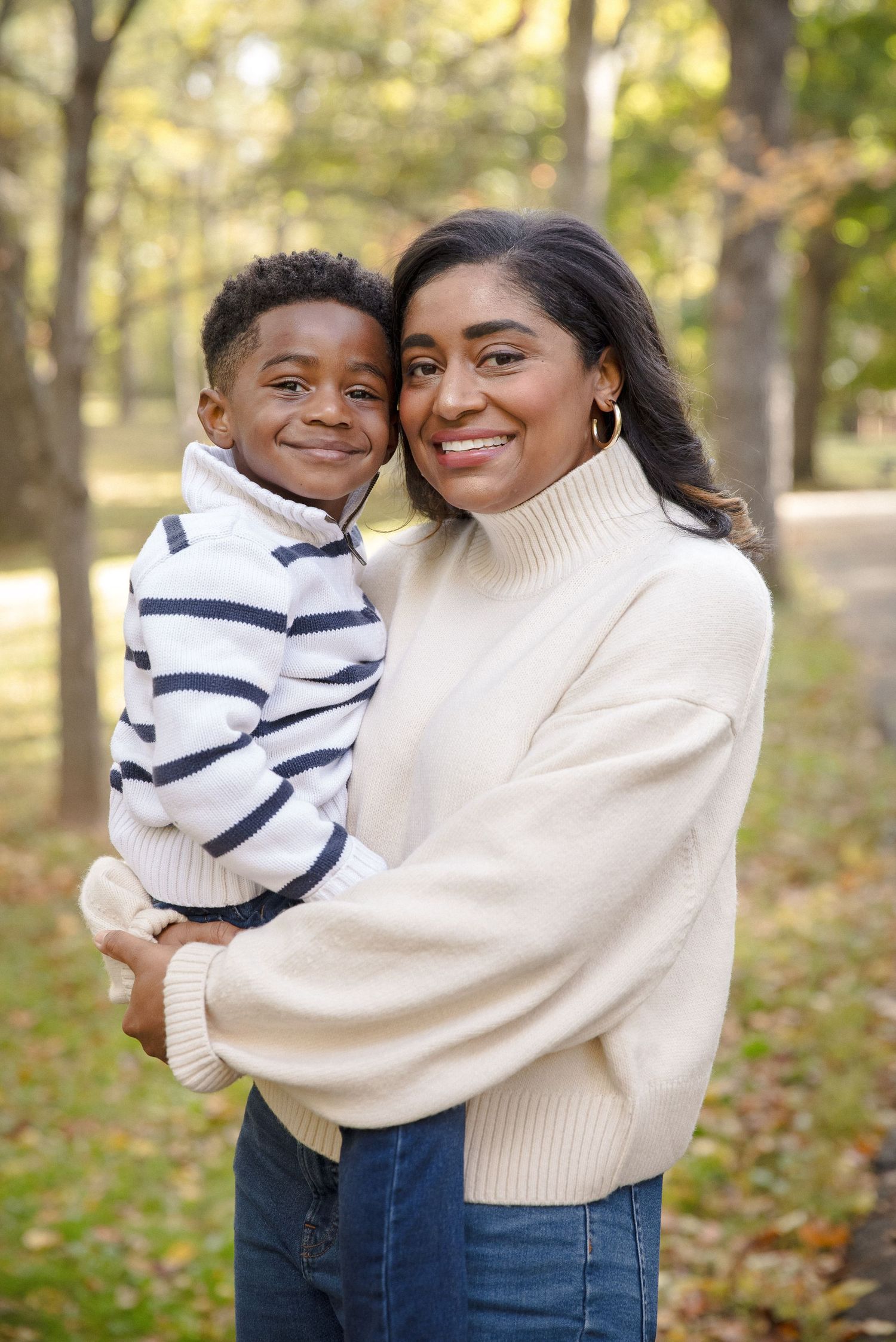 Smiling mother in cream sweater holds young son close in a sunlit autumn park.