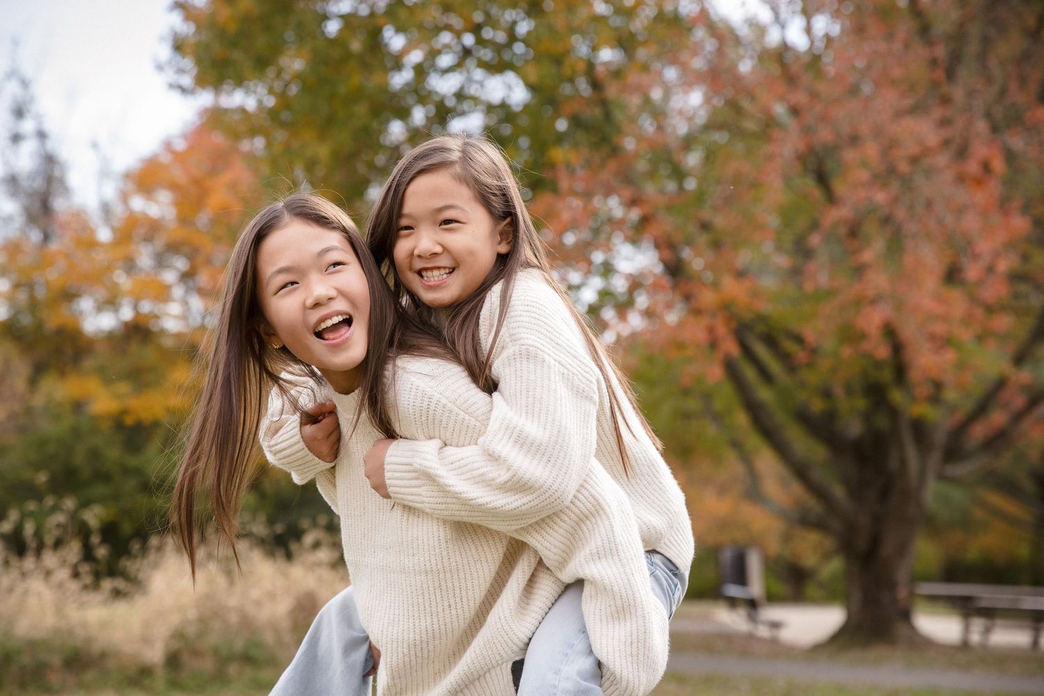 Two Asian sisters laughing joyfully as older sister gives younger one a piggyback ride in autumn park.