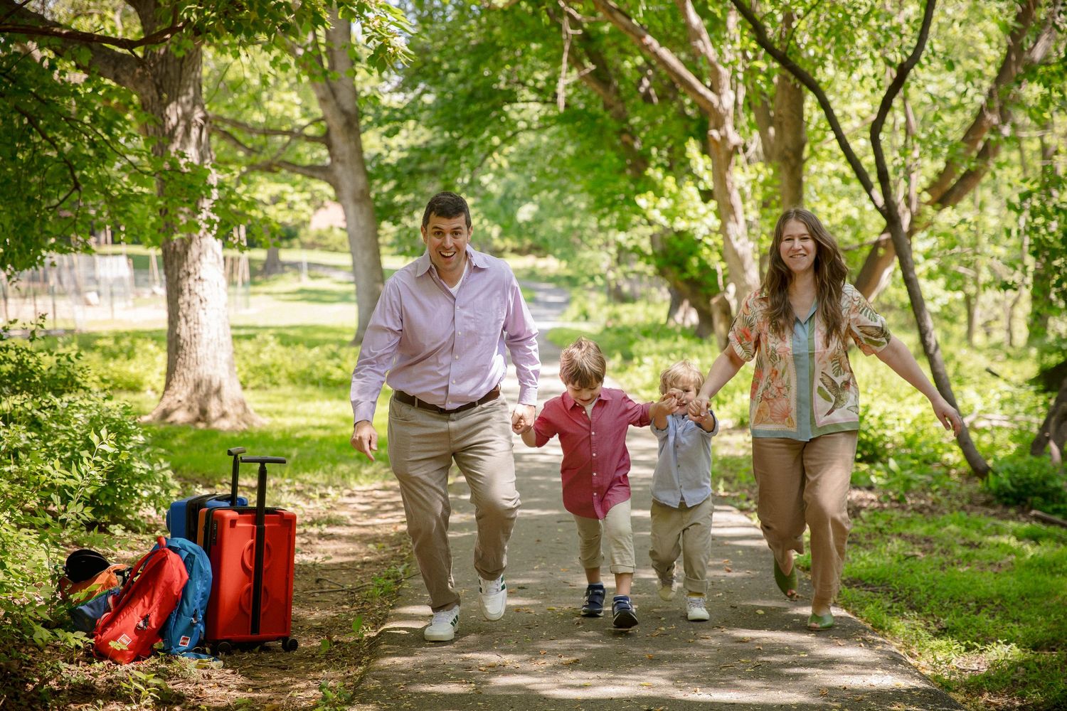 Family of four walking hand-in-hand on a sunny park path with red luggage nearby and lush green trees.