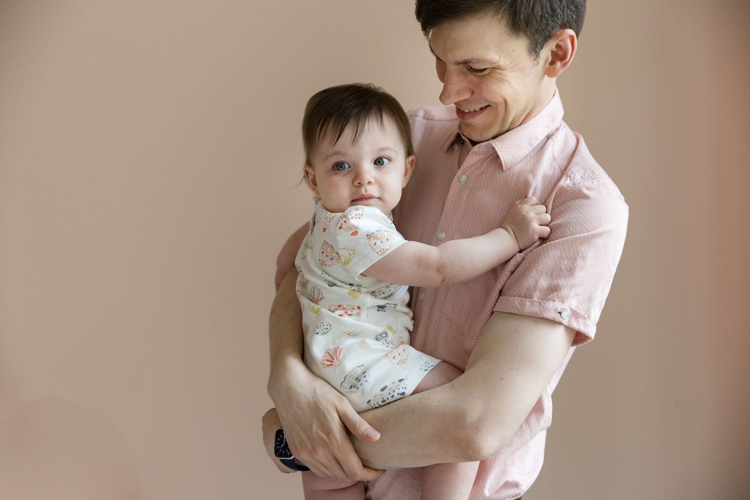 A smiling man in a pink shirt gently holds a baby dressed in a floral onesie against a beige background.