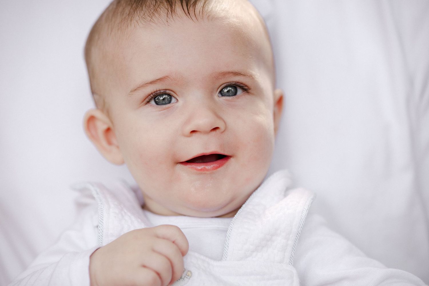 Smiling baby with blue eyes wearing a white outfit, lying on a white blanket.