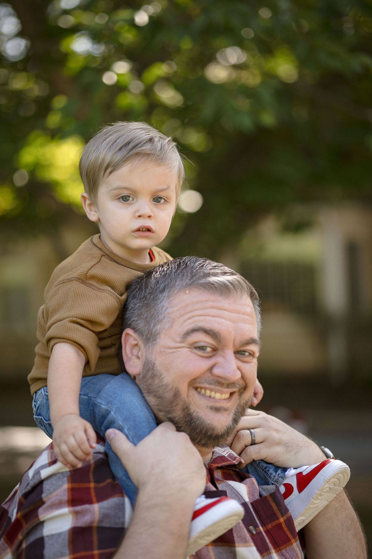 Happy father carrying toddler son on shoulders outdoors, smiling man with young blonde child in park setting.
