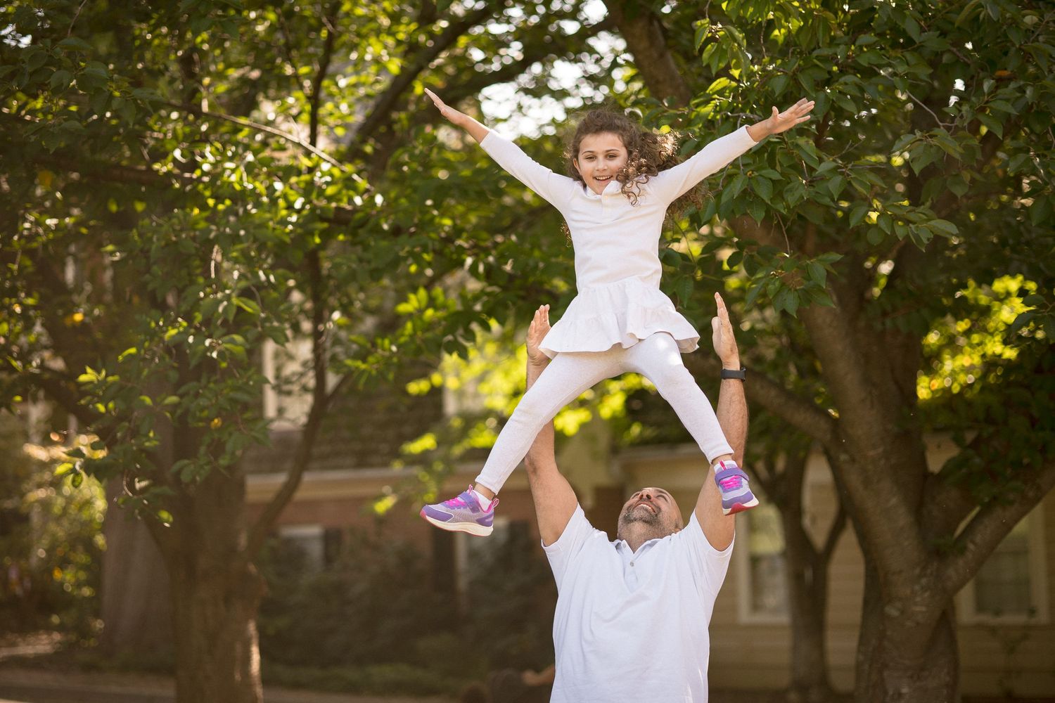 Man in white outfit lifting a young girl in white dress high above his head outdoors among green trees.