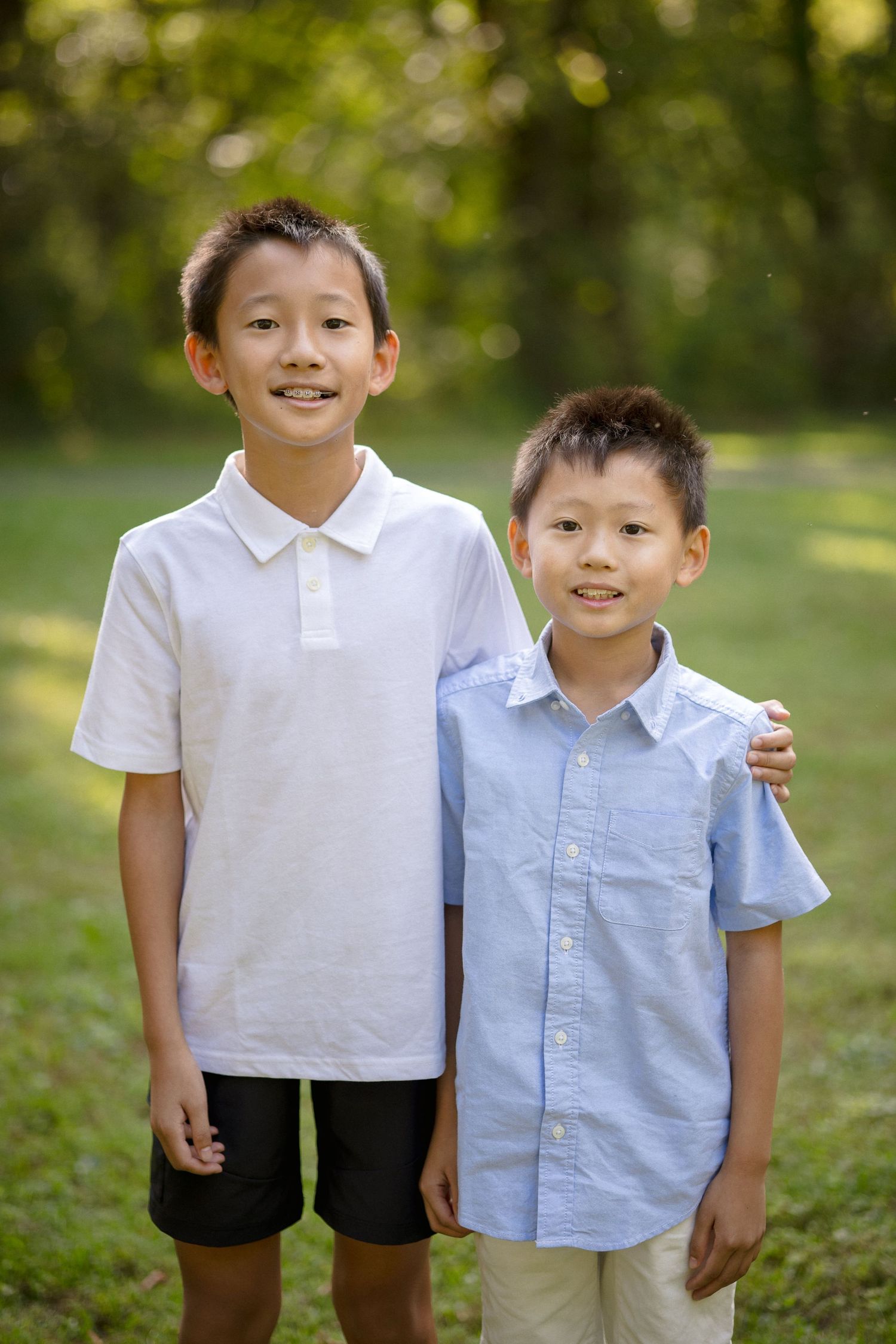 Two smiling Asian boys pose together outdoors in a green park, wearing casual button-up shirts.