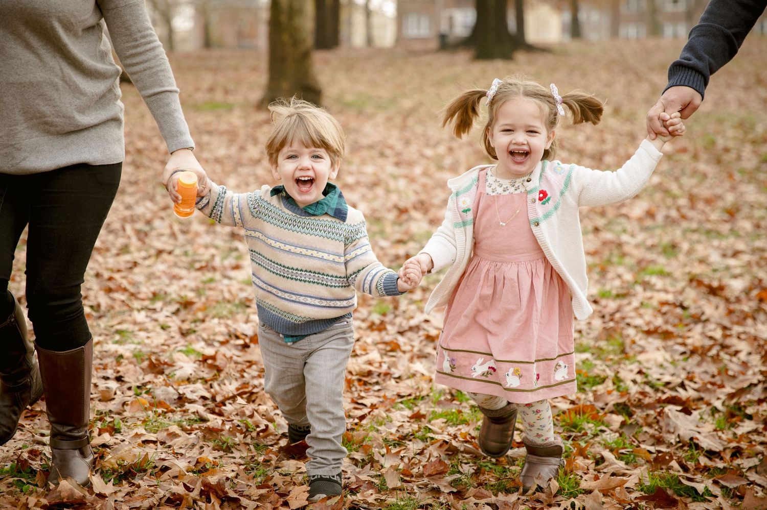 Two laughing toddlers hold hands with parents while playing in fall leaves.