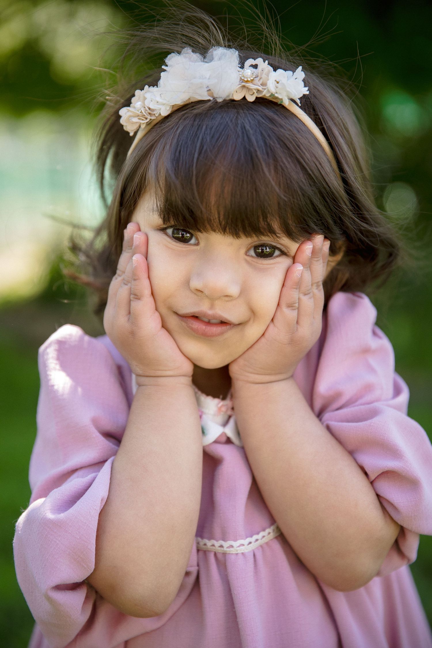Cute little girl in pink dress holding her cheeks with both hands wearing a floral headband outdoors.