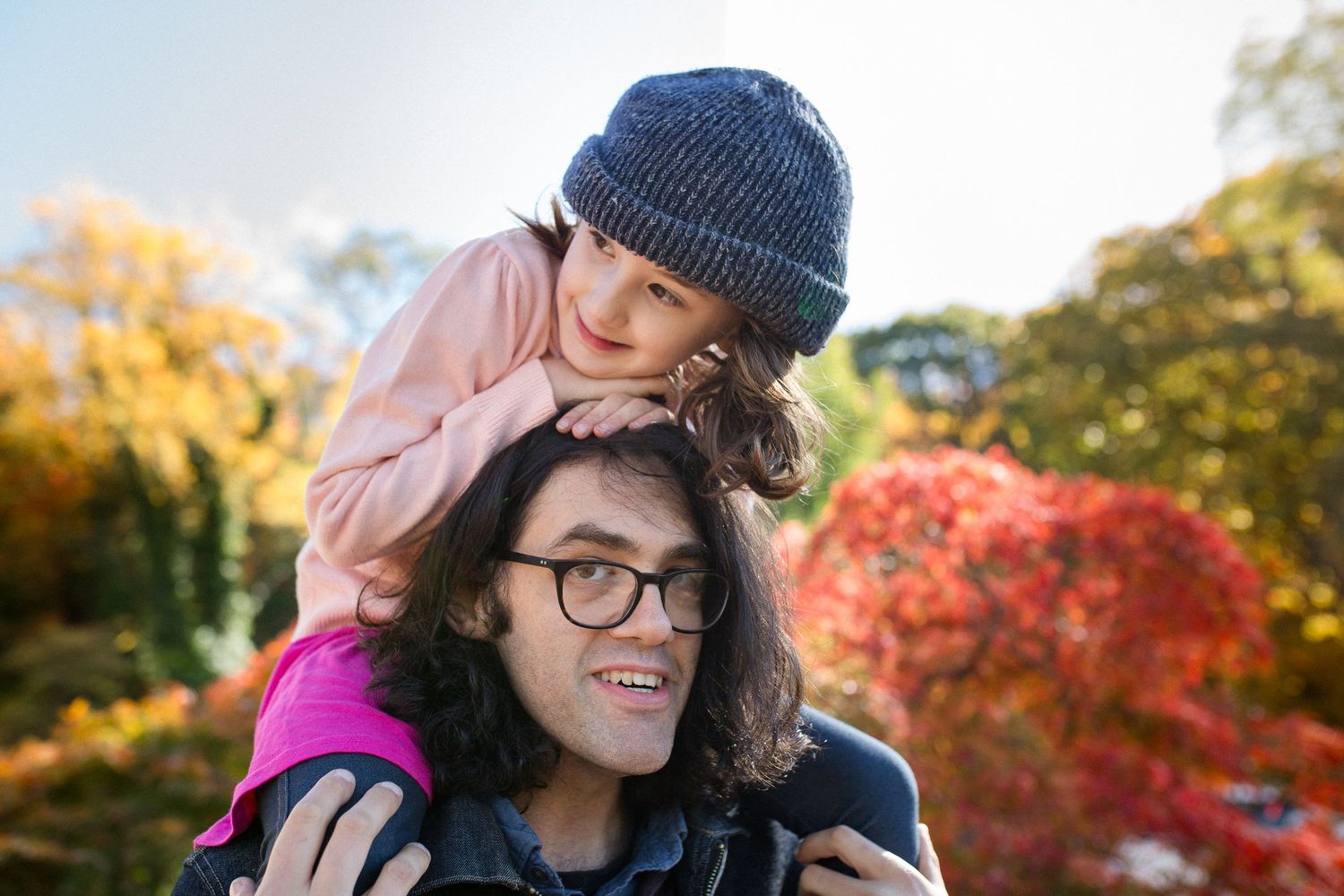 Young man with glasses carries smiling girl on shoulders outdoors among vibrant autumn foliage.