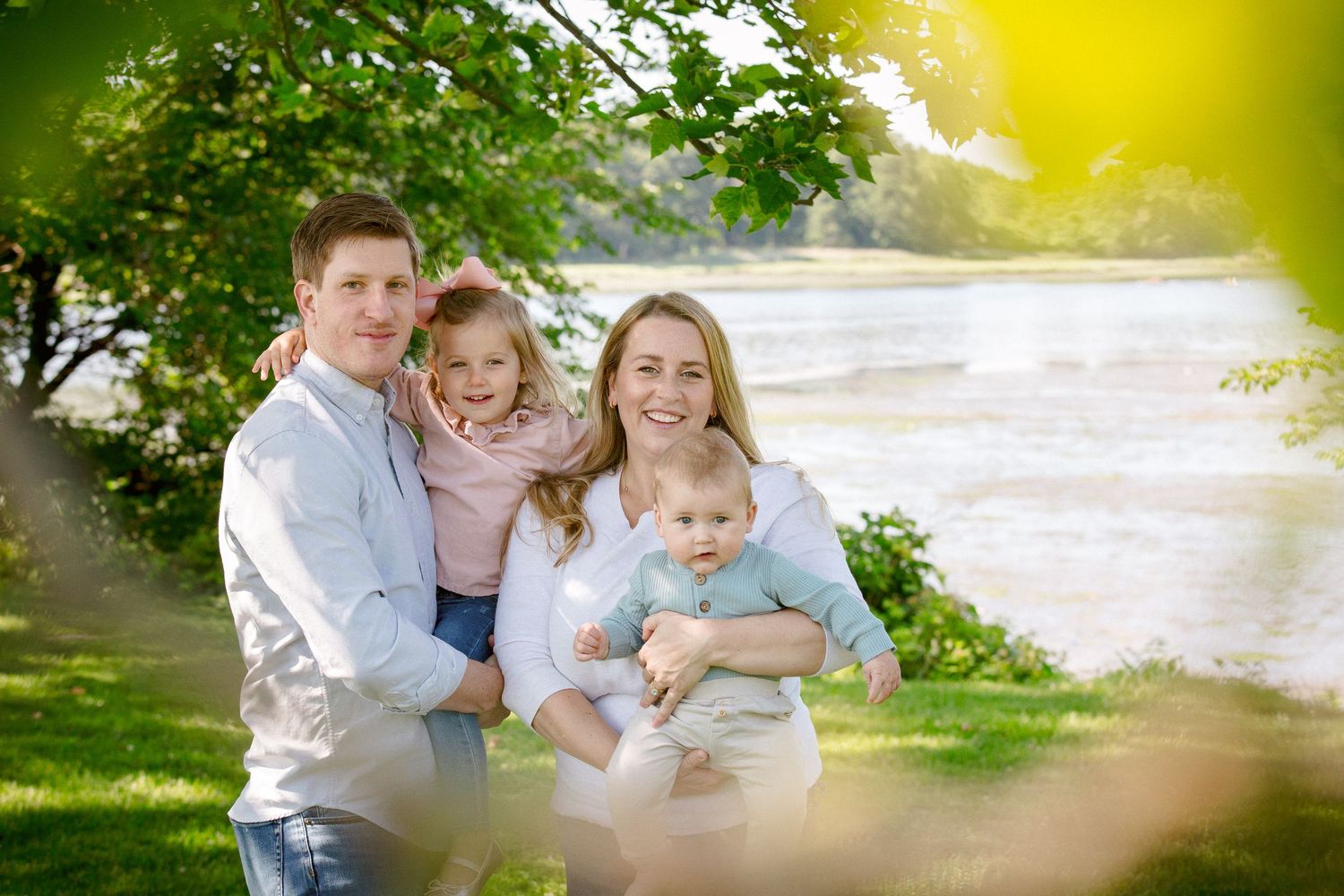 Happy family of four posing outdoors near a river under green trees on a sunny day.