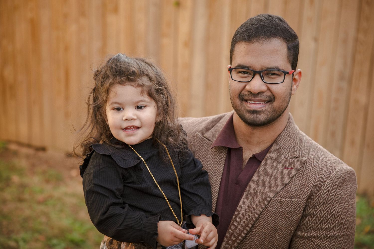 A man in a blazer holds a young curly-haired girl in a black dress outdoors near a wooden fence.