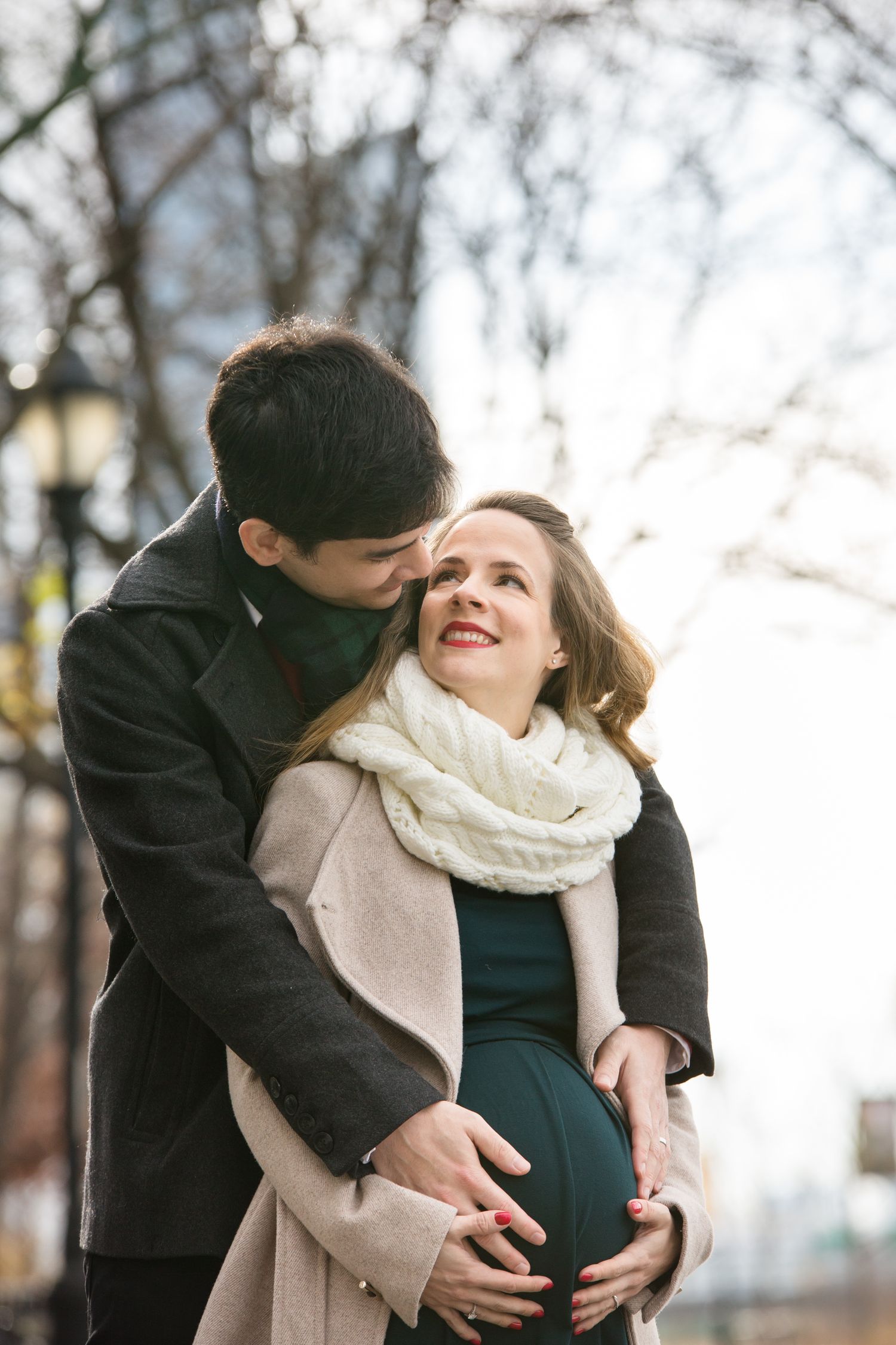 Expecting couple outdoors in winter, man kissing pregnant woman's head while holding her baby bump.