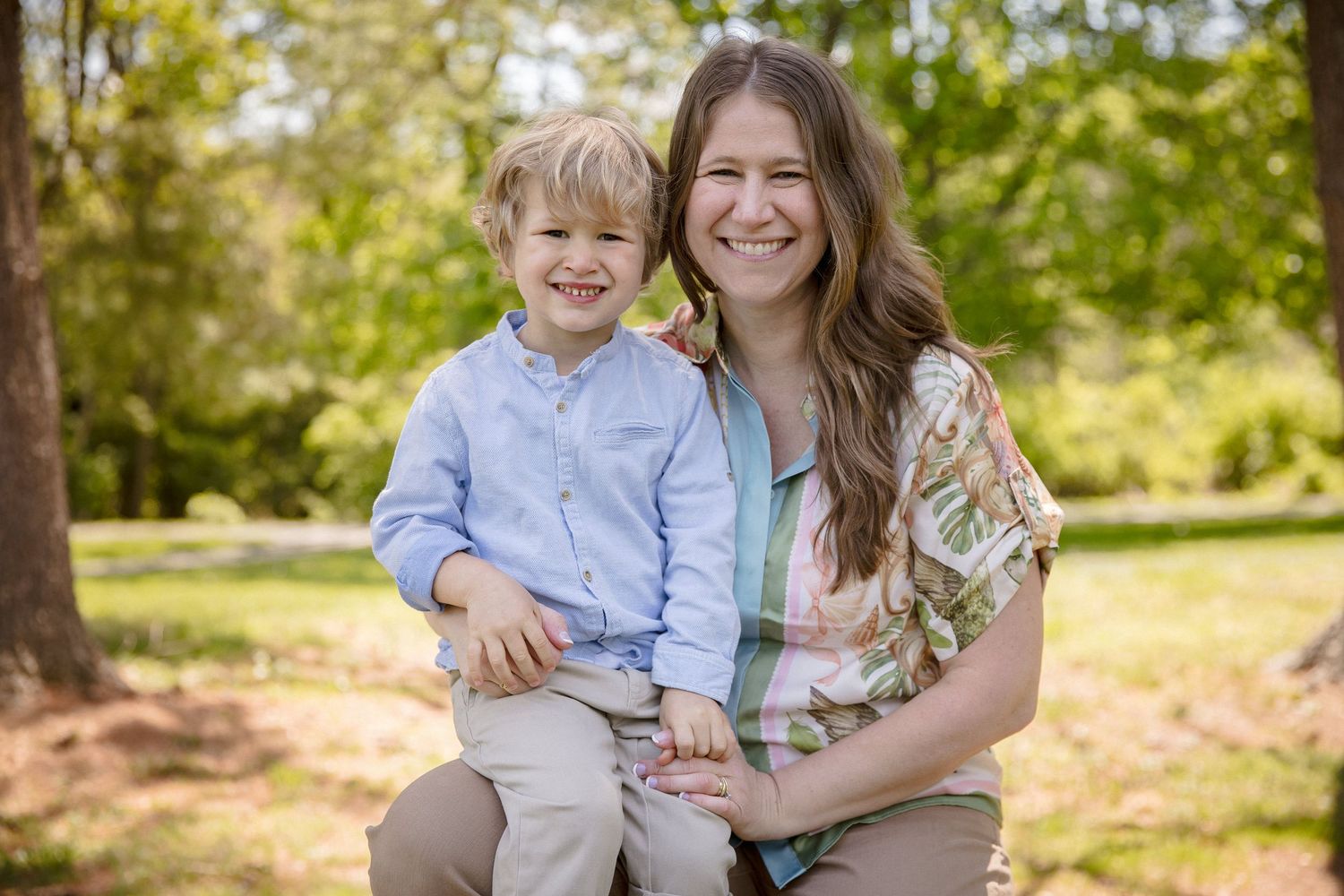 A woman and young blond boy smile together outdoors in a lush green park setting.