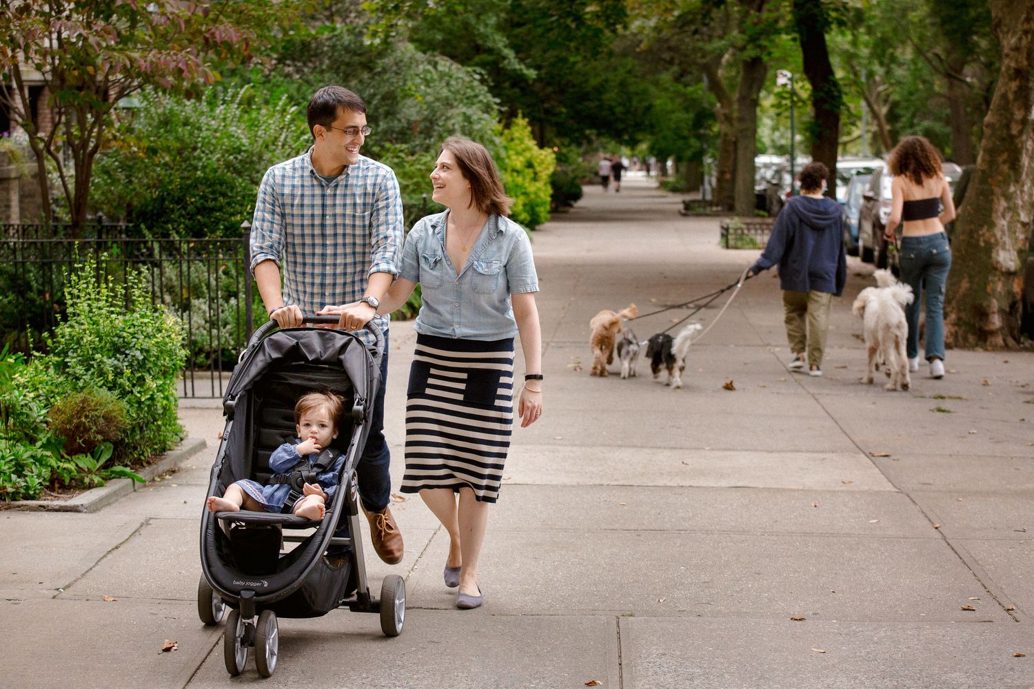 Young family strolling on city sidewalk with baby in black jogging stroller.