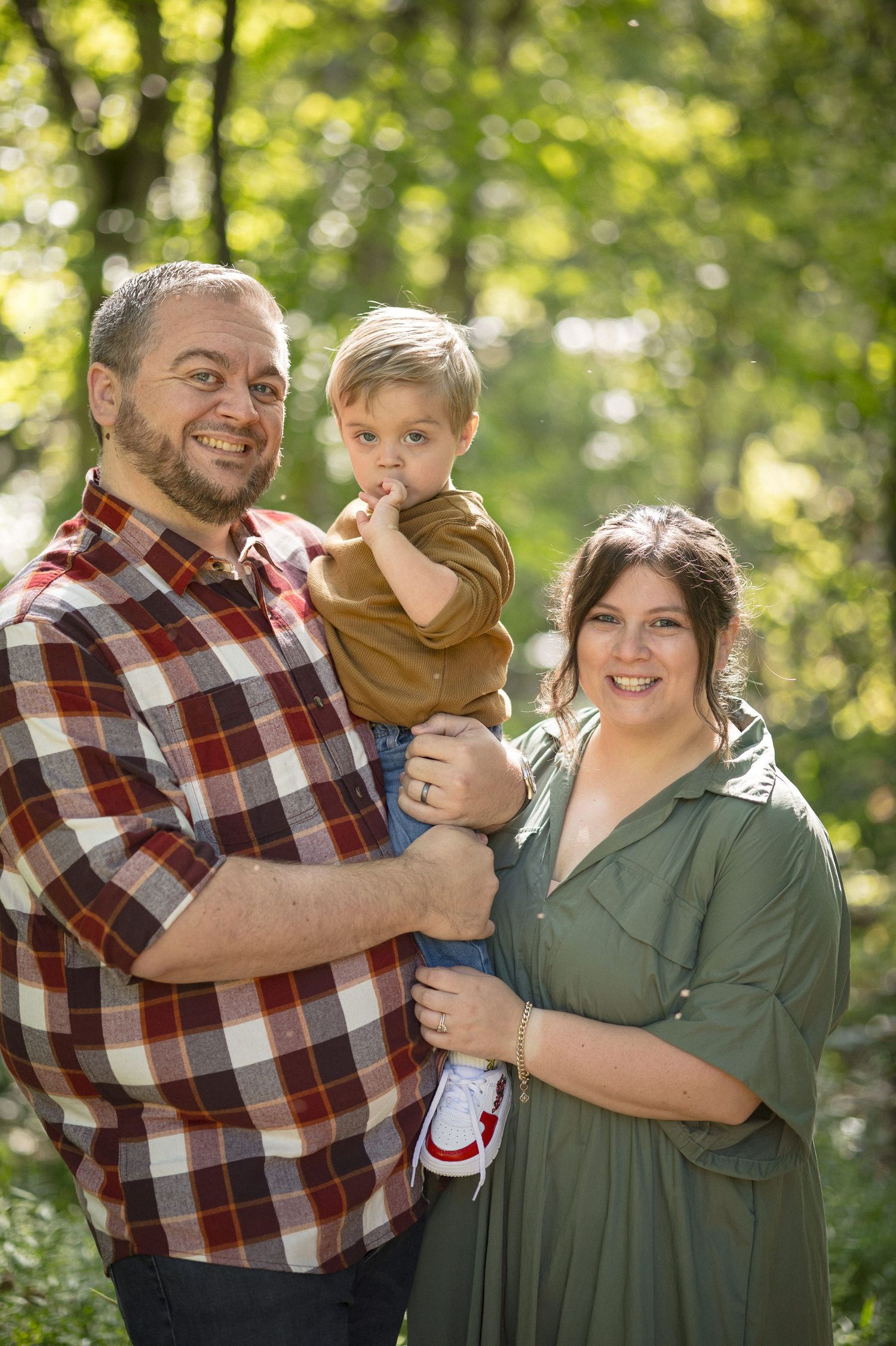 Happy family with toddler boy posing outdoors in green forest setting.