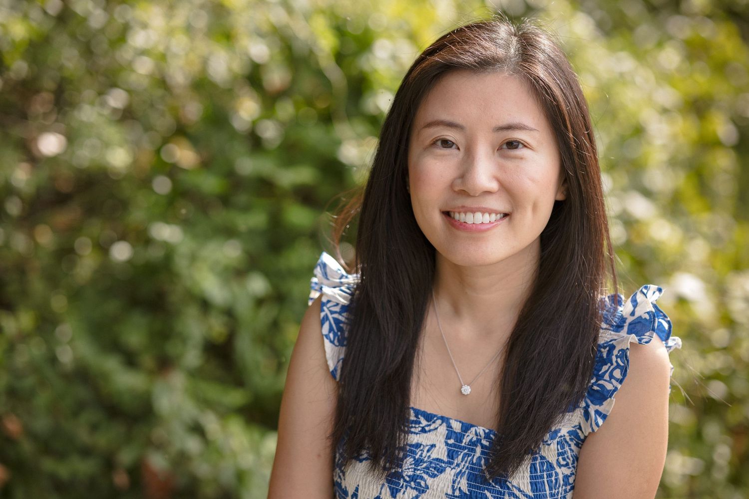 Smiling young woman with long dark hair wearing a blue floral top, posing outdoors with green foliage background.