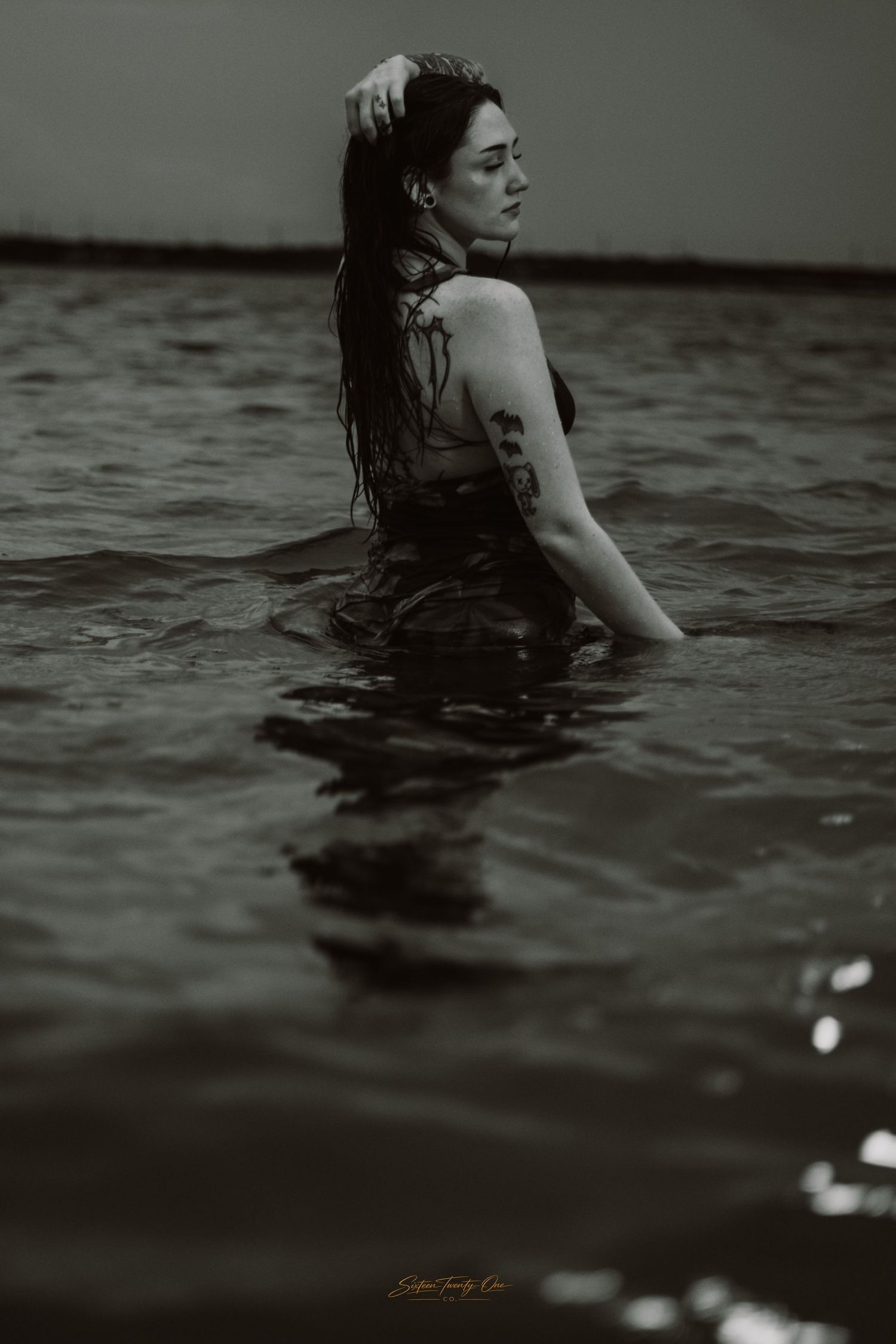 Black and white portrait of tattooed woman standing in water holding her hair.