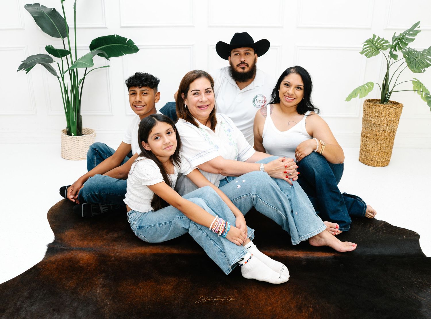 Happy family of five posing together in white shirts and jeans in a bright white studio with plants.