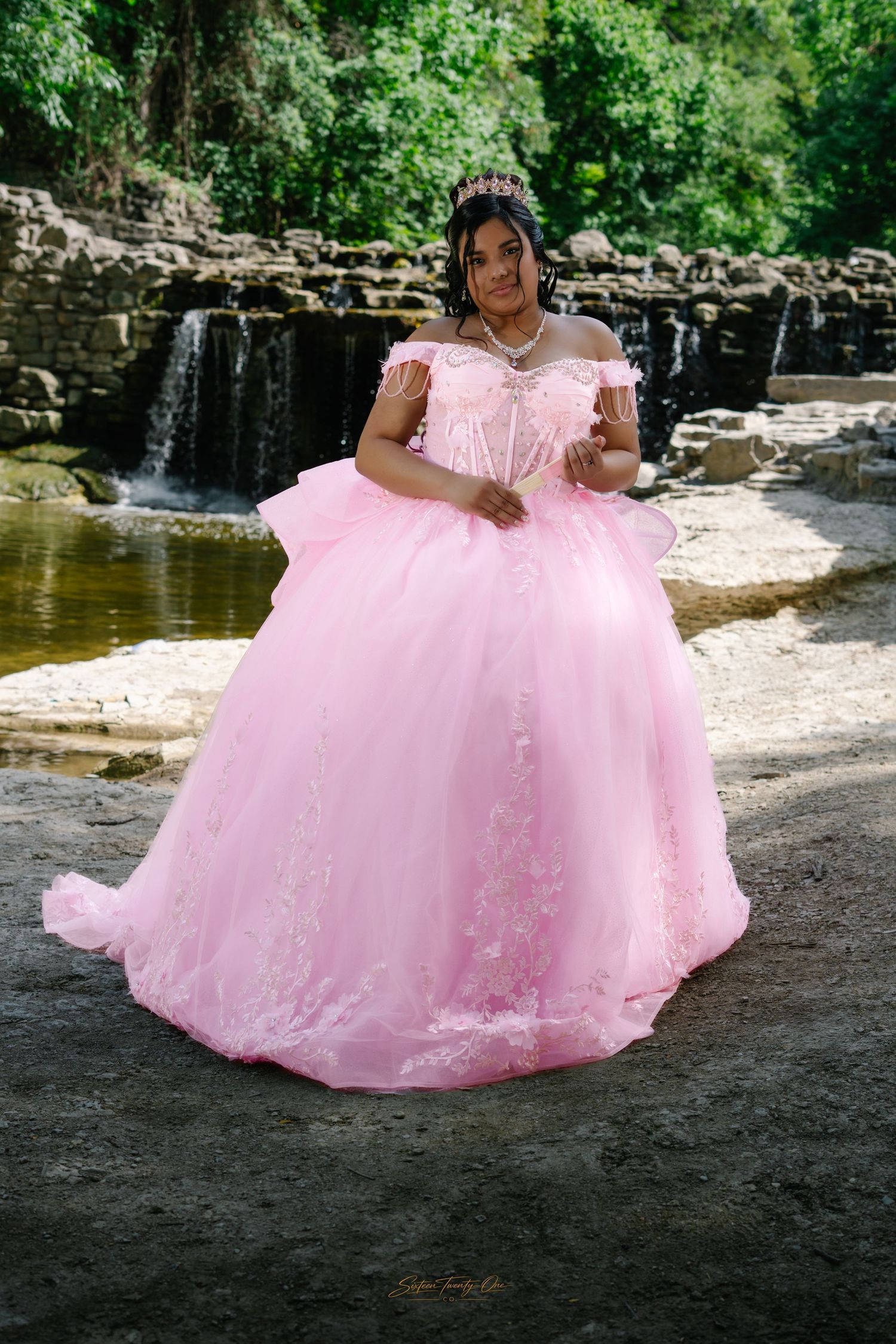 Young woman in a pink quinceañera ball gown with tiara posing near a waterfall.