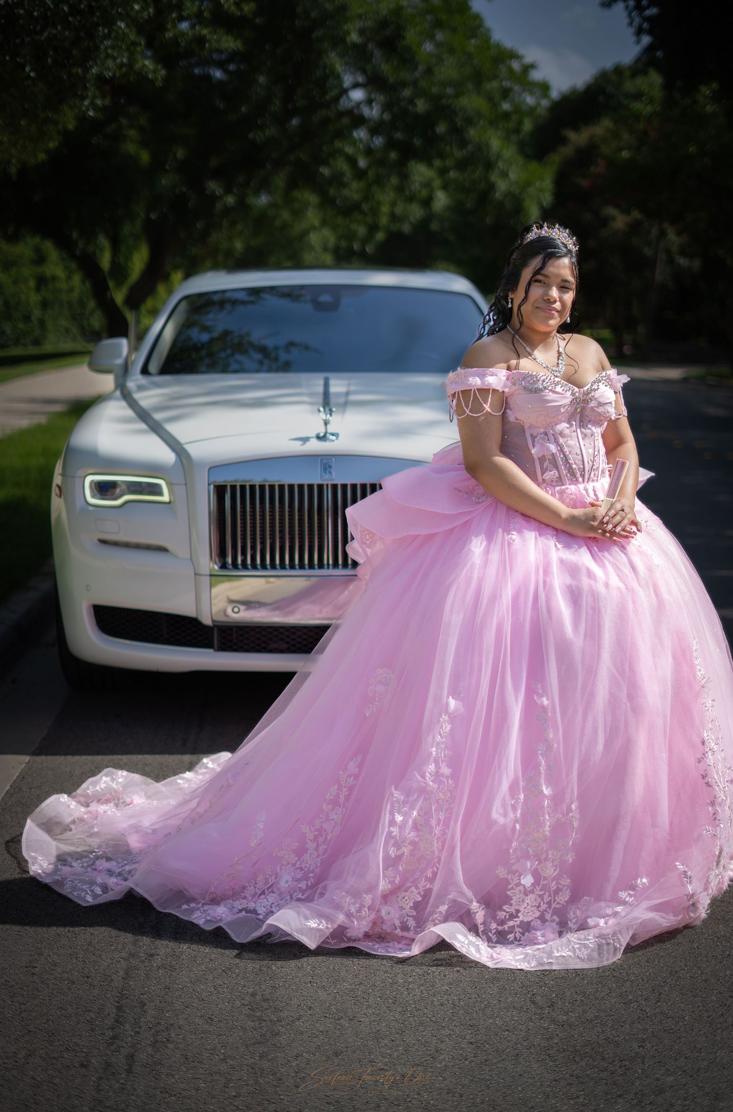 Young woman in pink quinceañera ball gown posing elegantly in front of a white Rolls-Royce outdoors.