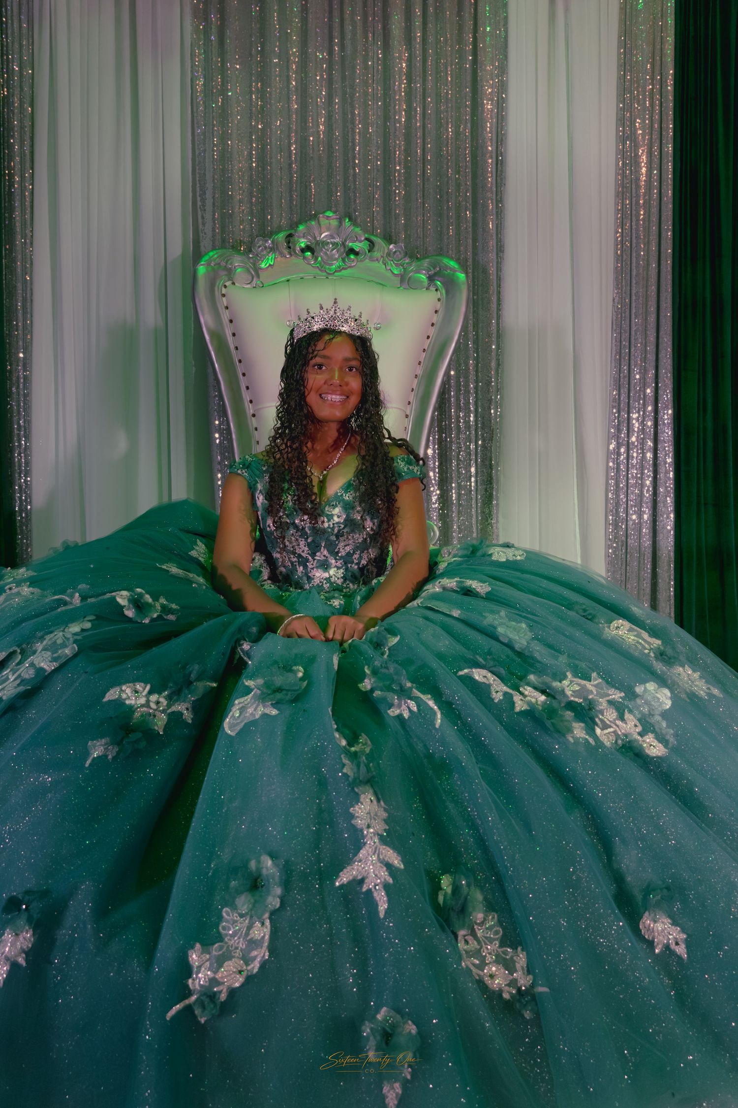 Smiling quinceañera in an emerald green ball gown and tiara sits on a decorative white throne.