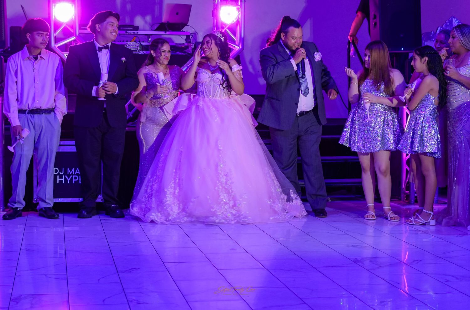 Quinceañera in pink ball gown celebrates on a white dance floor surrounded by guests and DJ lights.