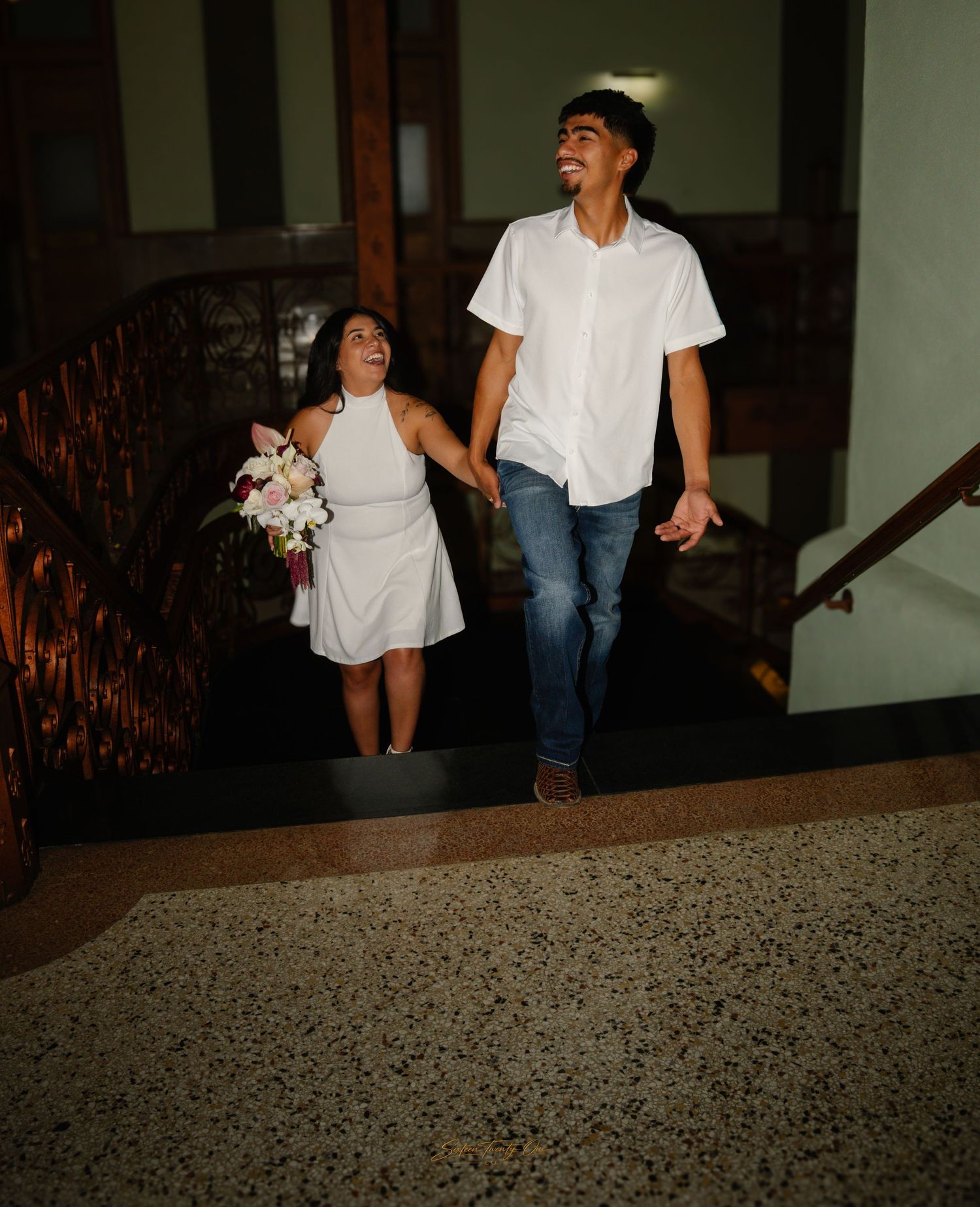 Joyful newlywed couple in white outfits walking up grand staircase, bride holding floral bouquet.