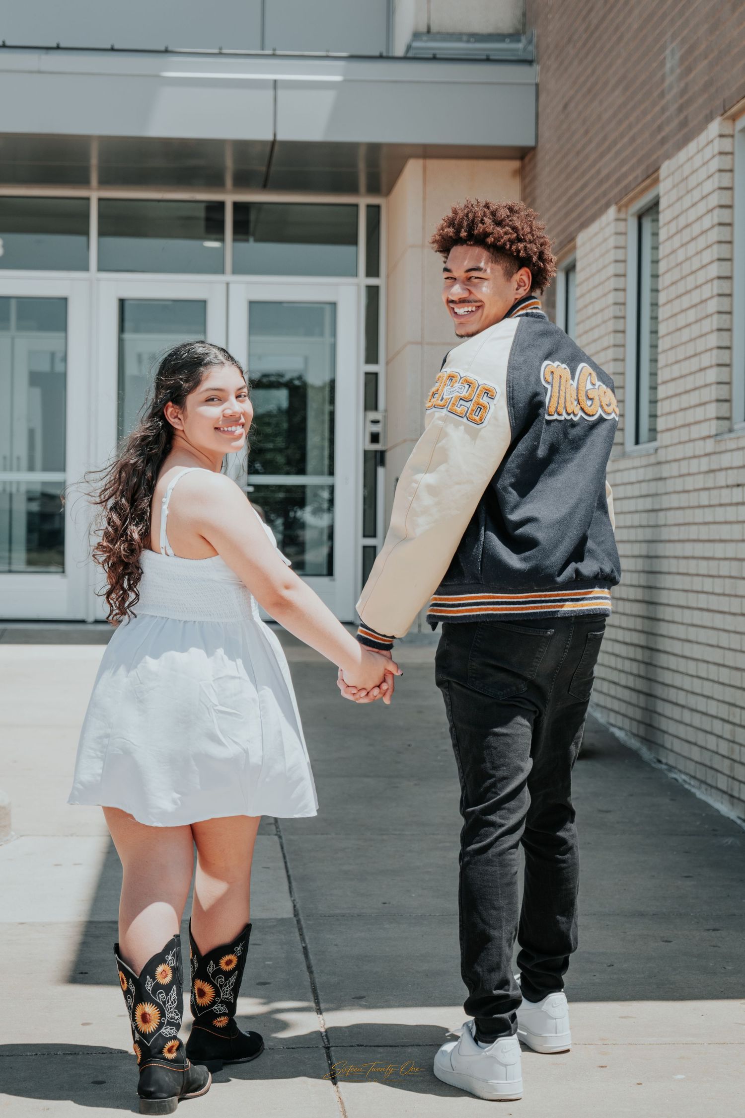 Smiling couple holding hands outside school, girl in white dress with cowboy boots, boy in varsity jacket.