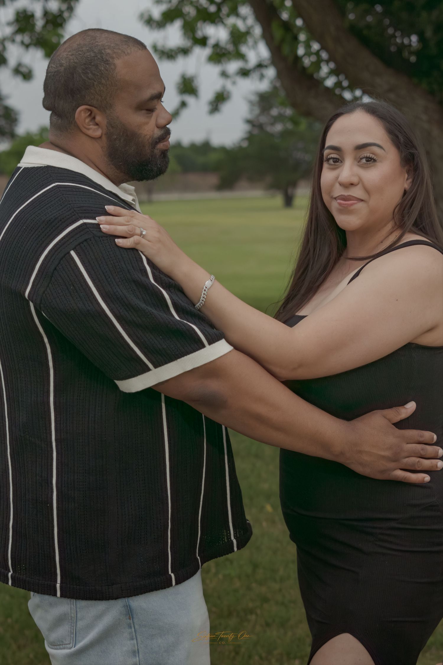 Couple posing outdoors under tree, man in black striped polo and woman in black dress embracing.