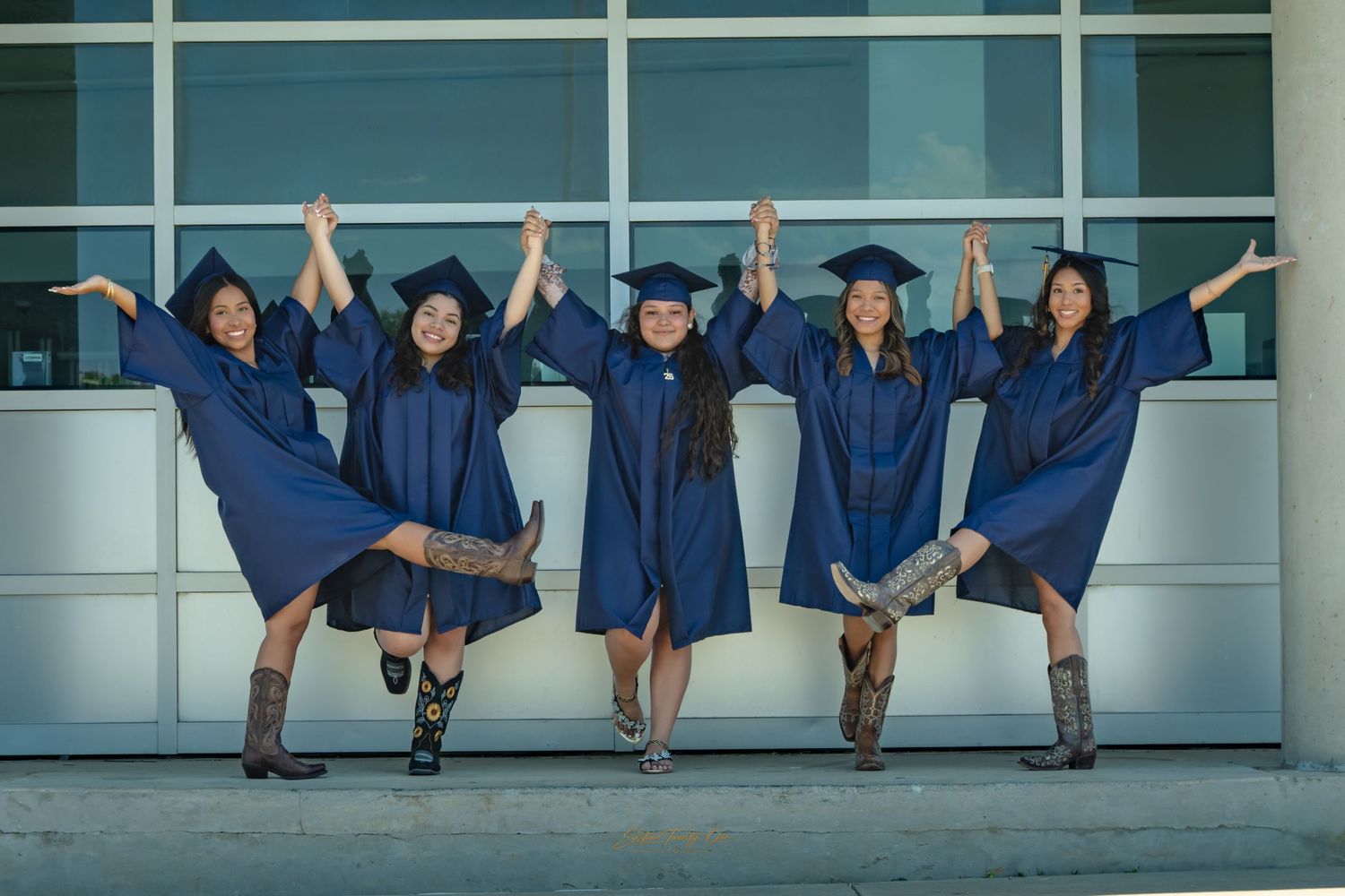 Five female graduates in blue caps and gowns joyfully holding hands raised outside a modern building.