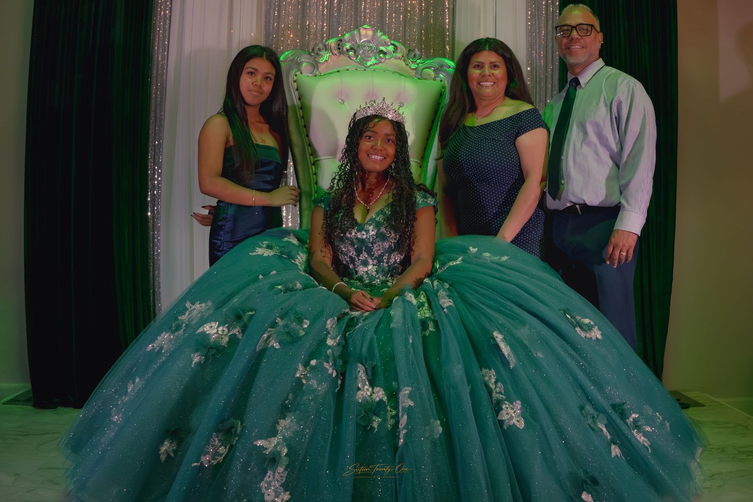 Quinceañera in green ball gown and tiara poses with family on a decorated throne chair backdrop.